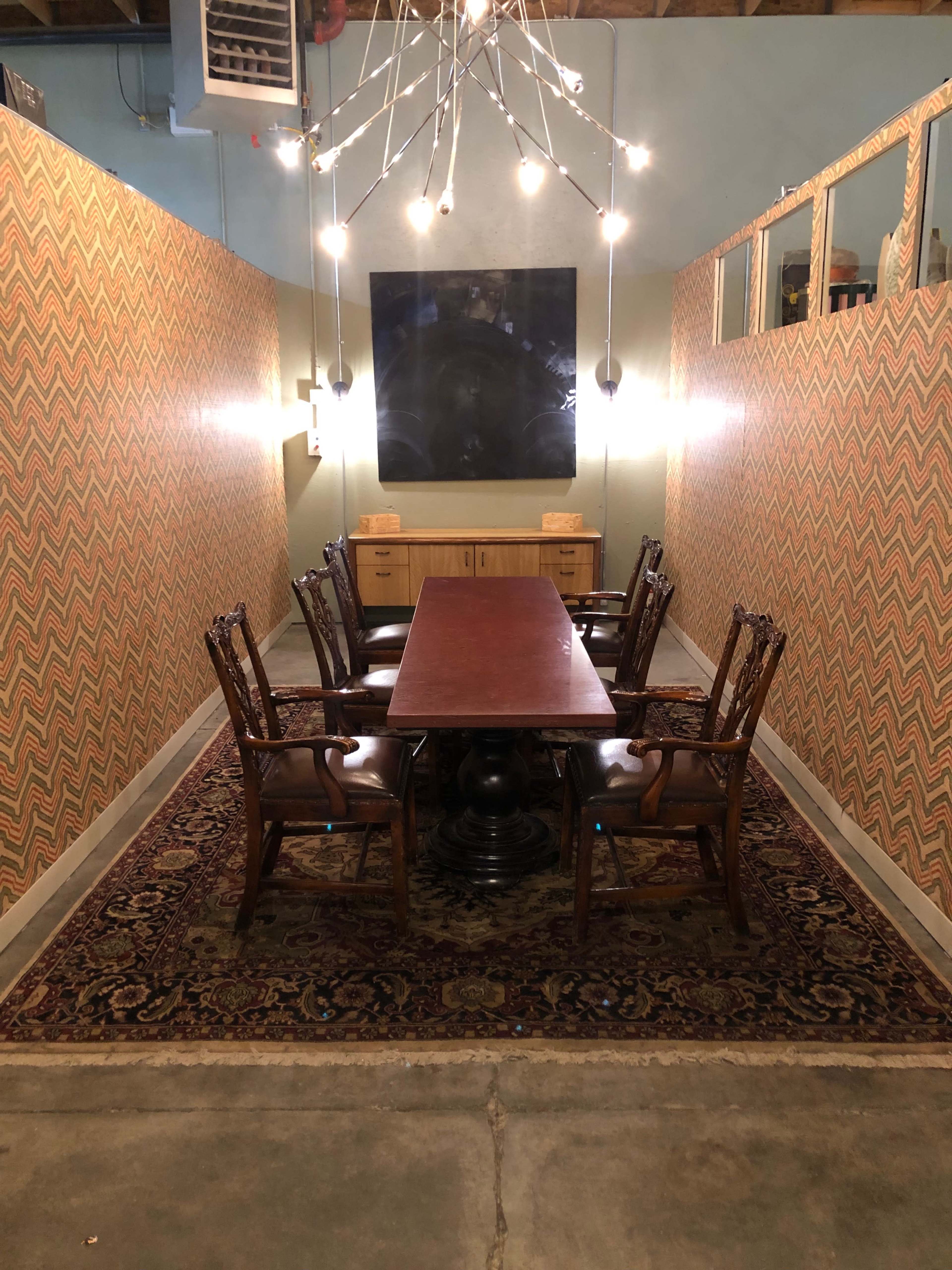A long wooden table surrounded by six ornate chairs is set in a brightly lit meeting room with patterned walls and a large dark artwork at the far end.