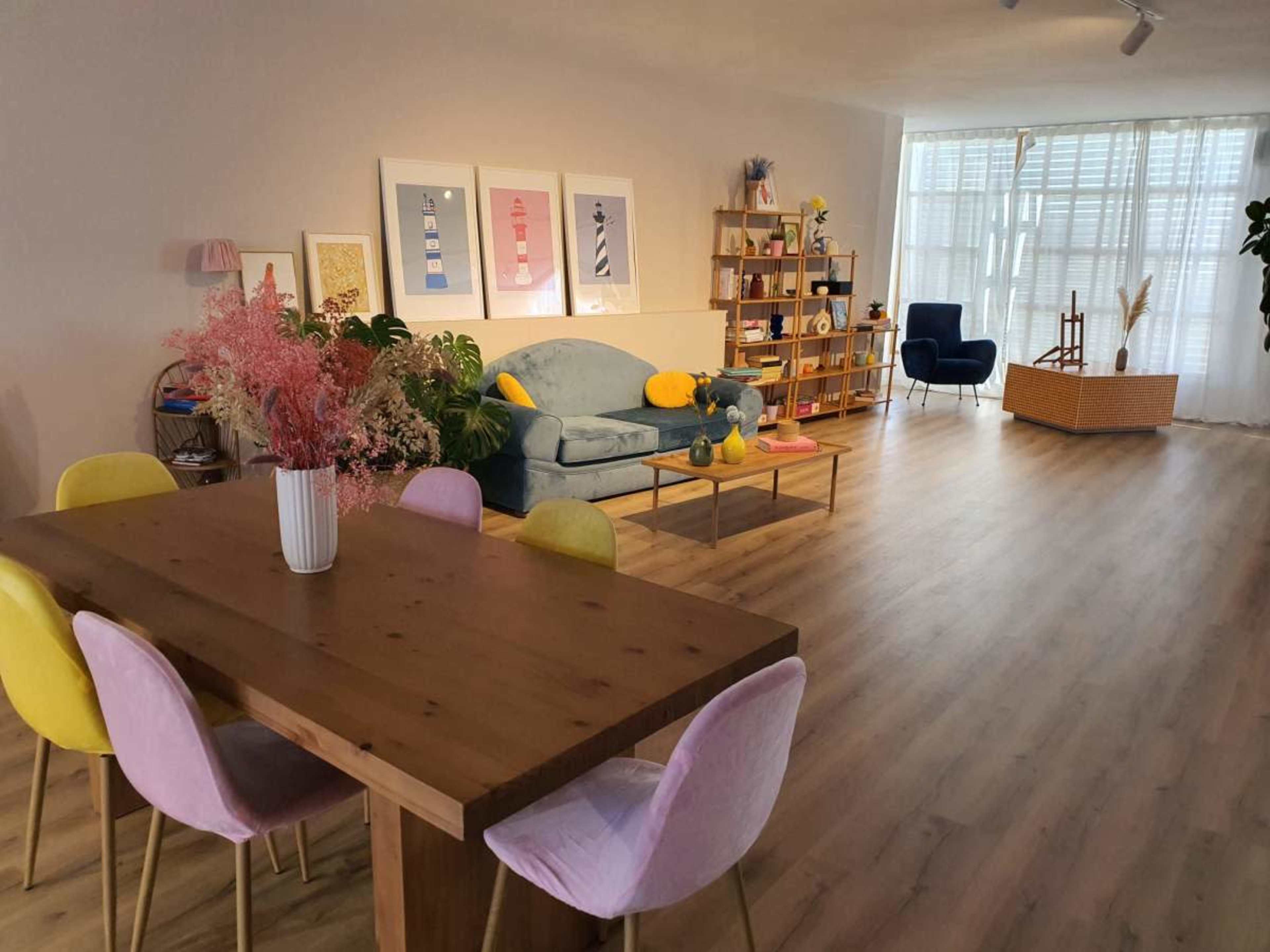 The image shows a modern living space featuring a wooden dining table with colorful chairs, a light blue couch, a bookshelf, and large windows allowing natural light.
