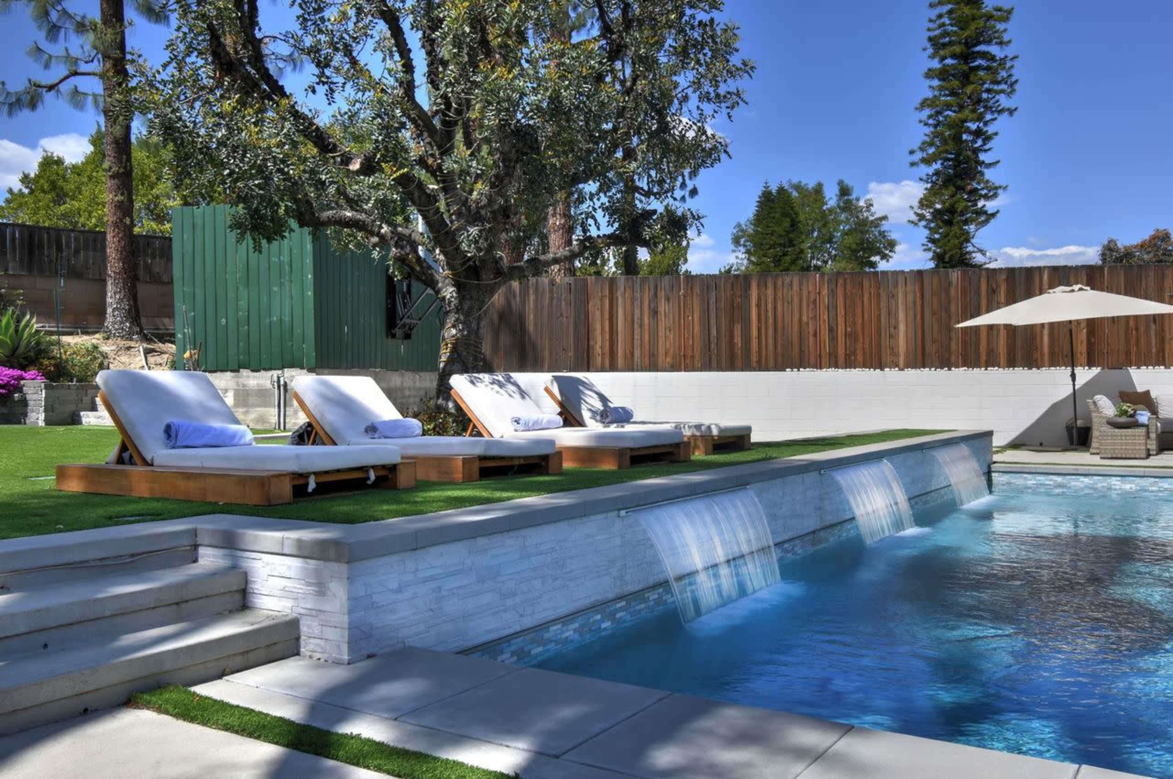 The image depicts a backyard pool area featuring three lounge chairs by the water, a stone wall with three water features, and a neatly landscaped garden under a clear blue sky.
