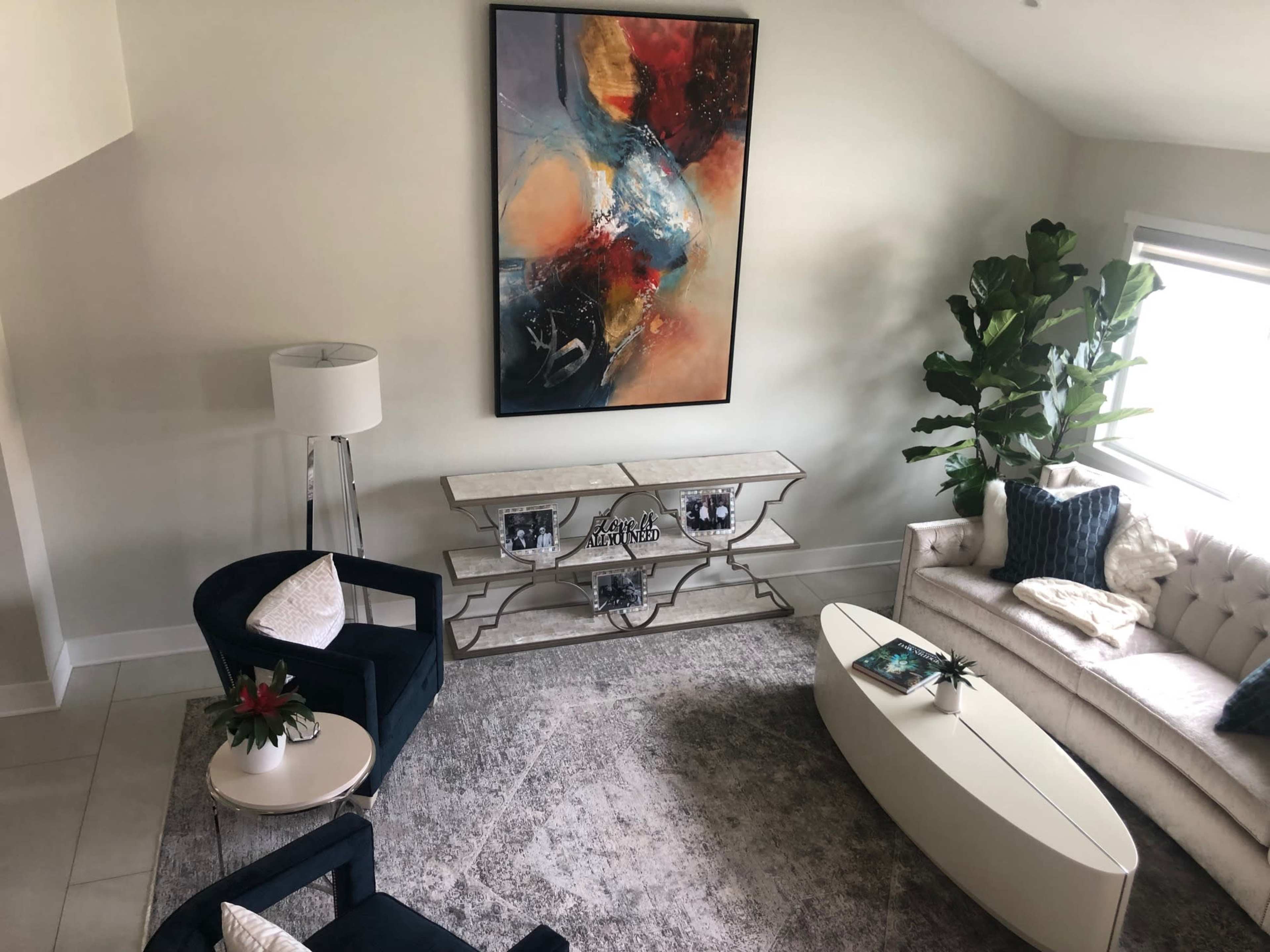 The image shows a modern living room featuring a large abstract painting, a white sofa, two dark blue chairs, a decorative table, and a tall plant by the window.