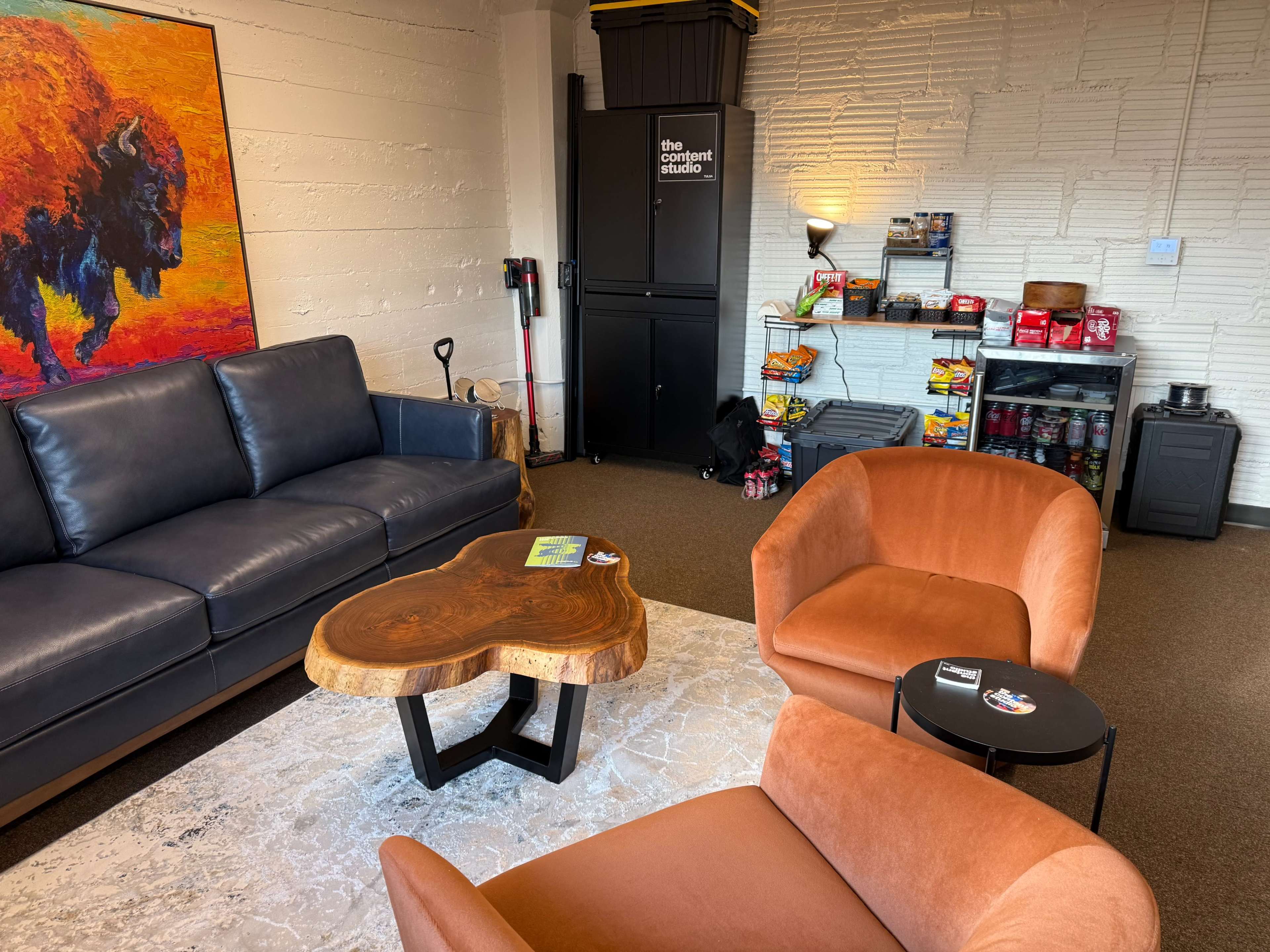 The image shows a cozy lounge area with a dark blue sofa, two orange armchairs, a wooden coffee table, and a small food and drink station in the background.