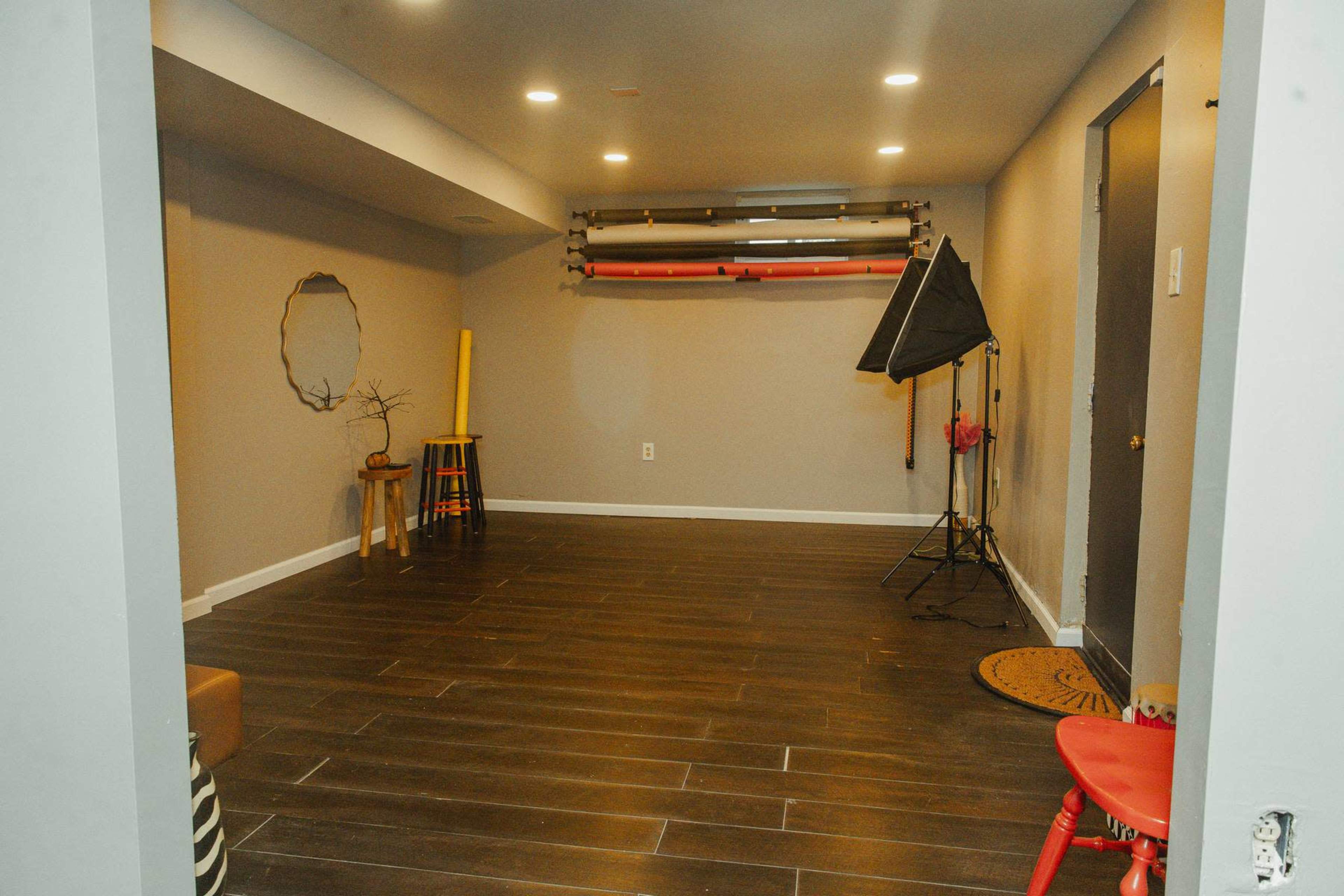 The image shows a spacious, empty room with dark wooden floors, grey walls, and studio lighting equipment positioned against one wall.