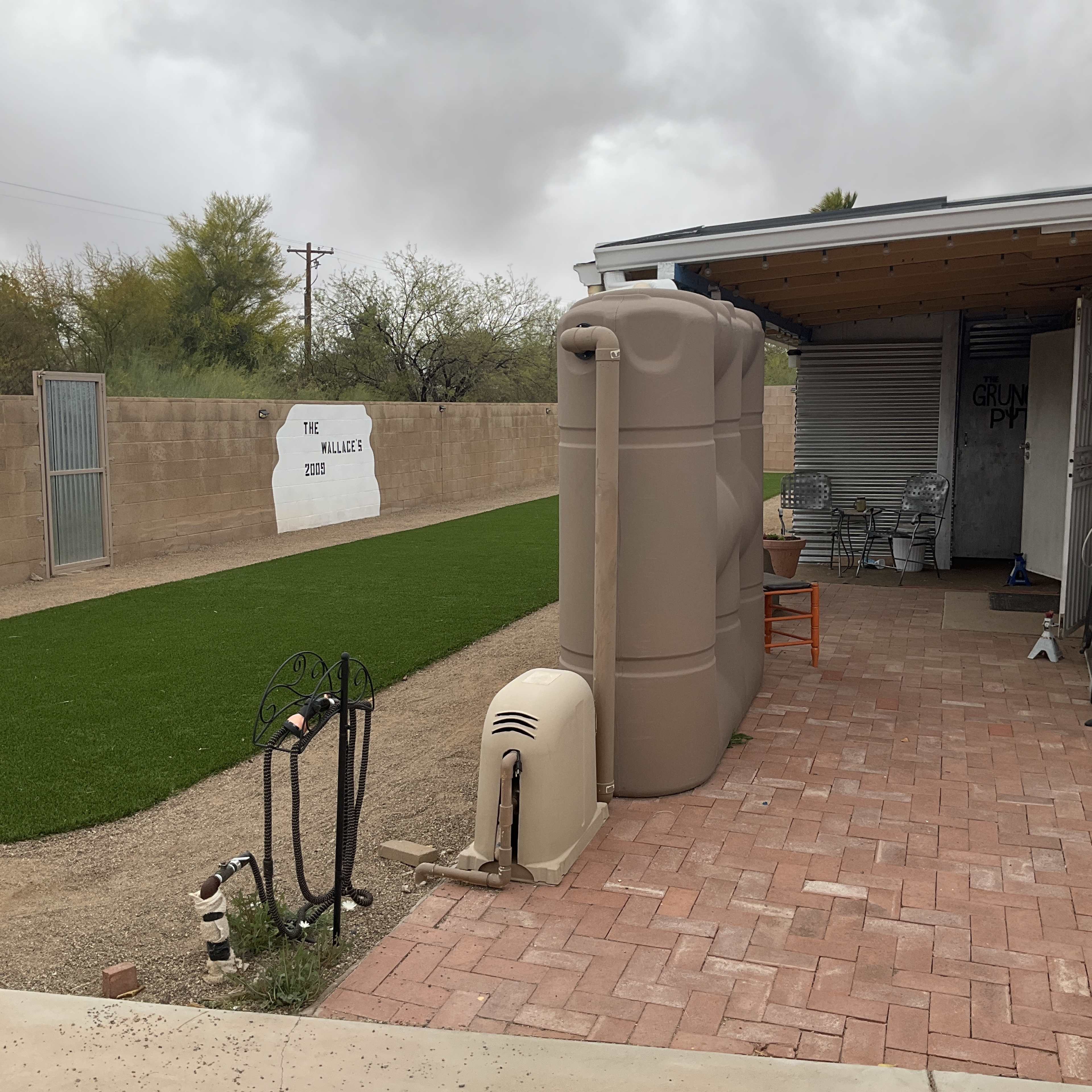 The image shows a backyard area with synthetic grass, a beige water storage tank, and a paved patio adjacent to a wall displaying an art piece.