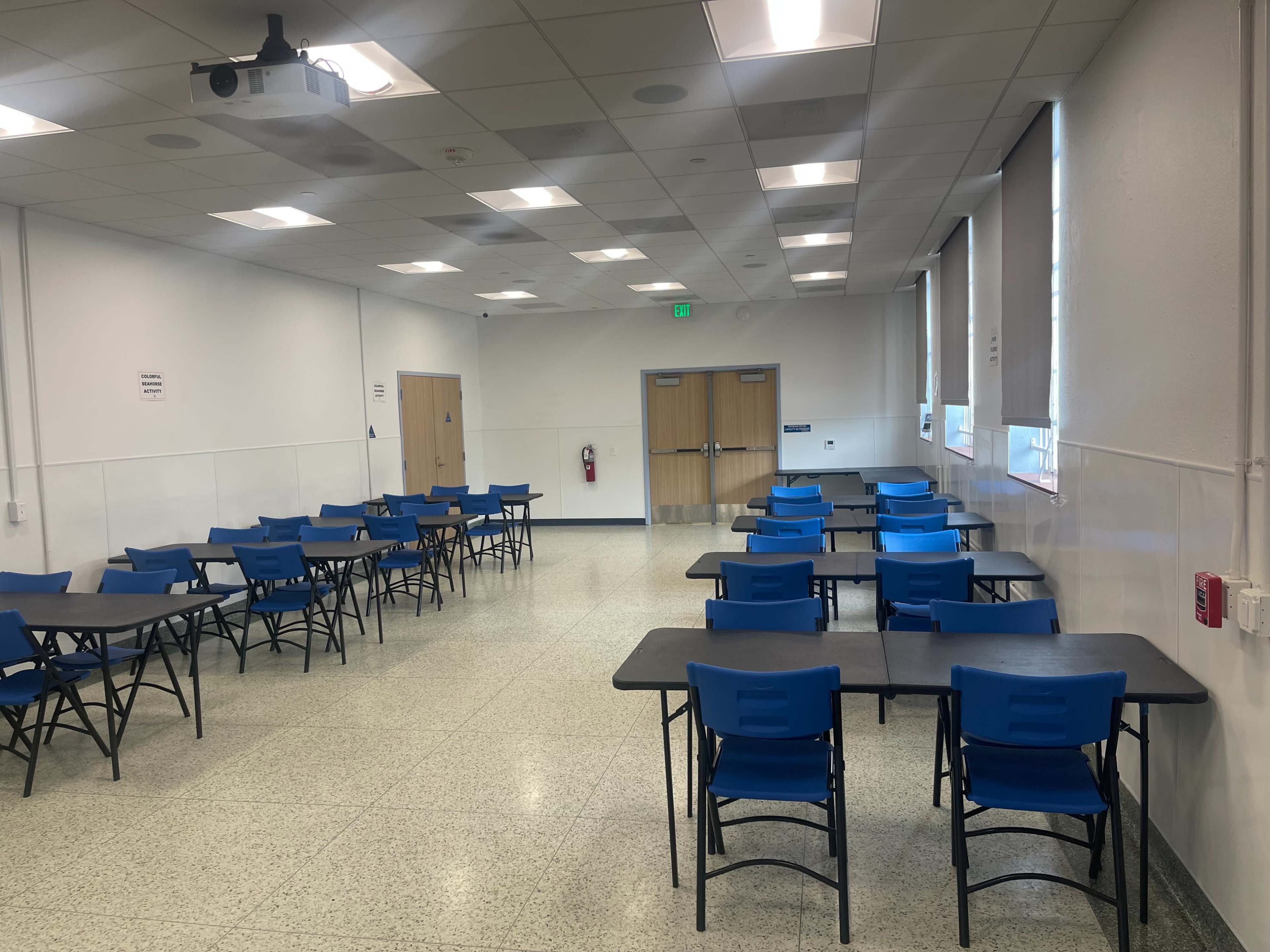 The image shows a classroom or meeting space filled with blue tables and chairs arranged in rows, with a projector mounted on the ceiling.