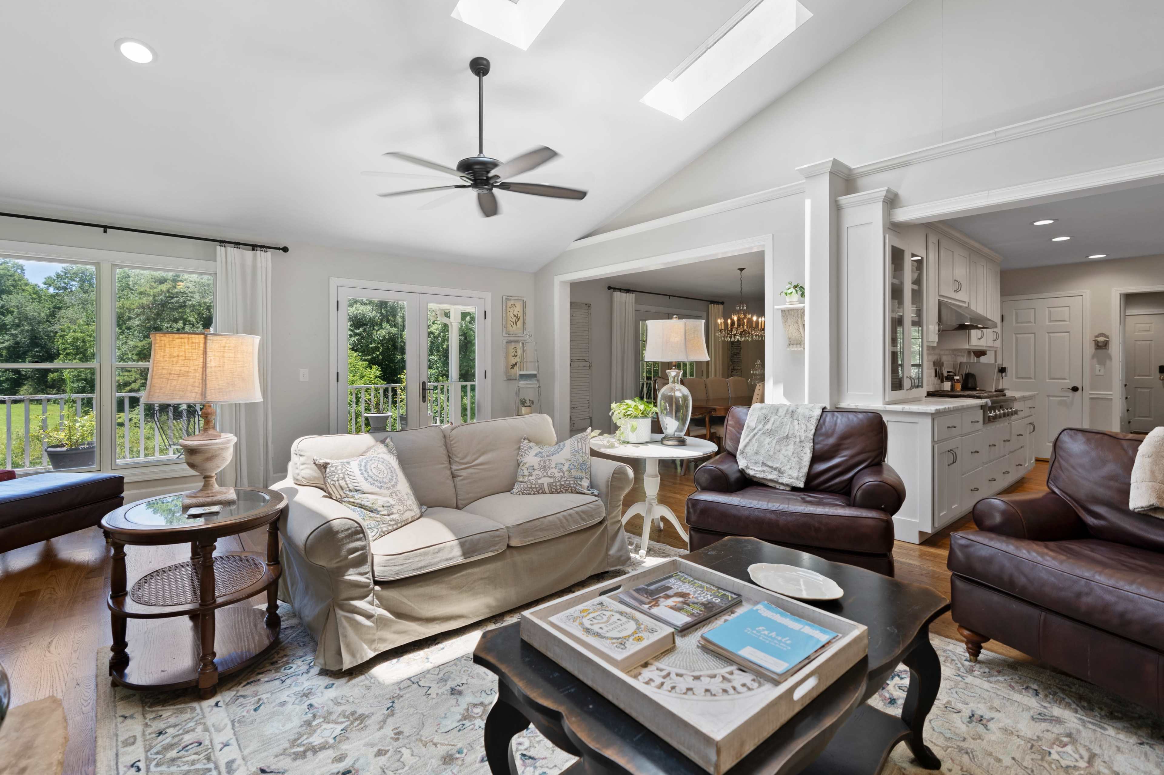 A bright, spacious living room features a light-colored sofa, dark leather chairs, and large windows with a view of greenery.