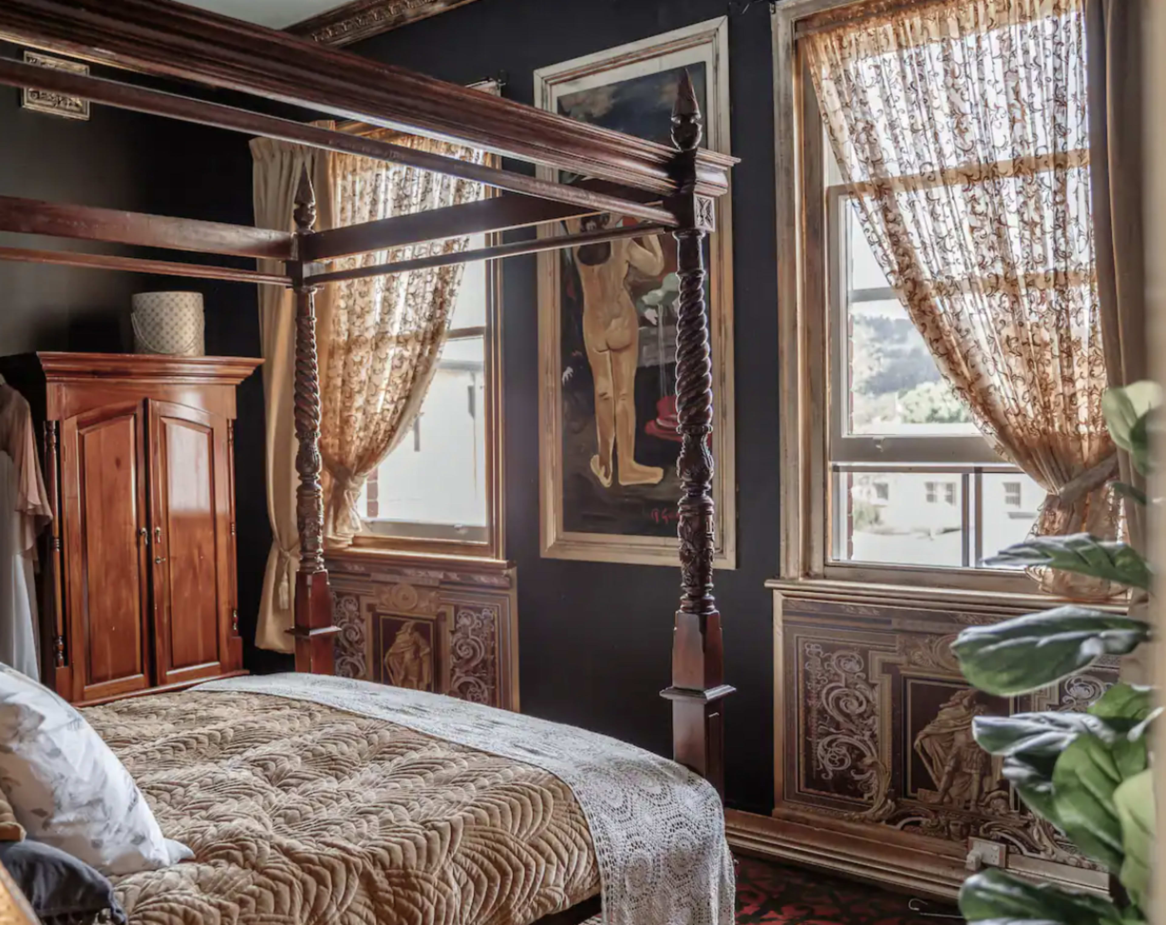 The room features a four-poster bed, wooden wardrobe, and large windows with patterned curtains, complemented by framed artwork and decorative wall panels.