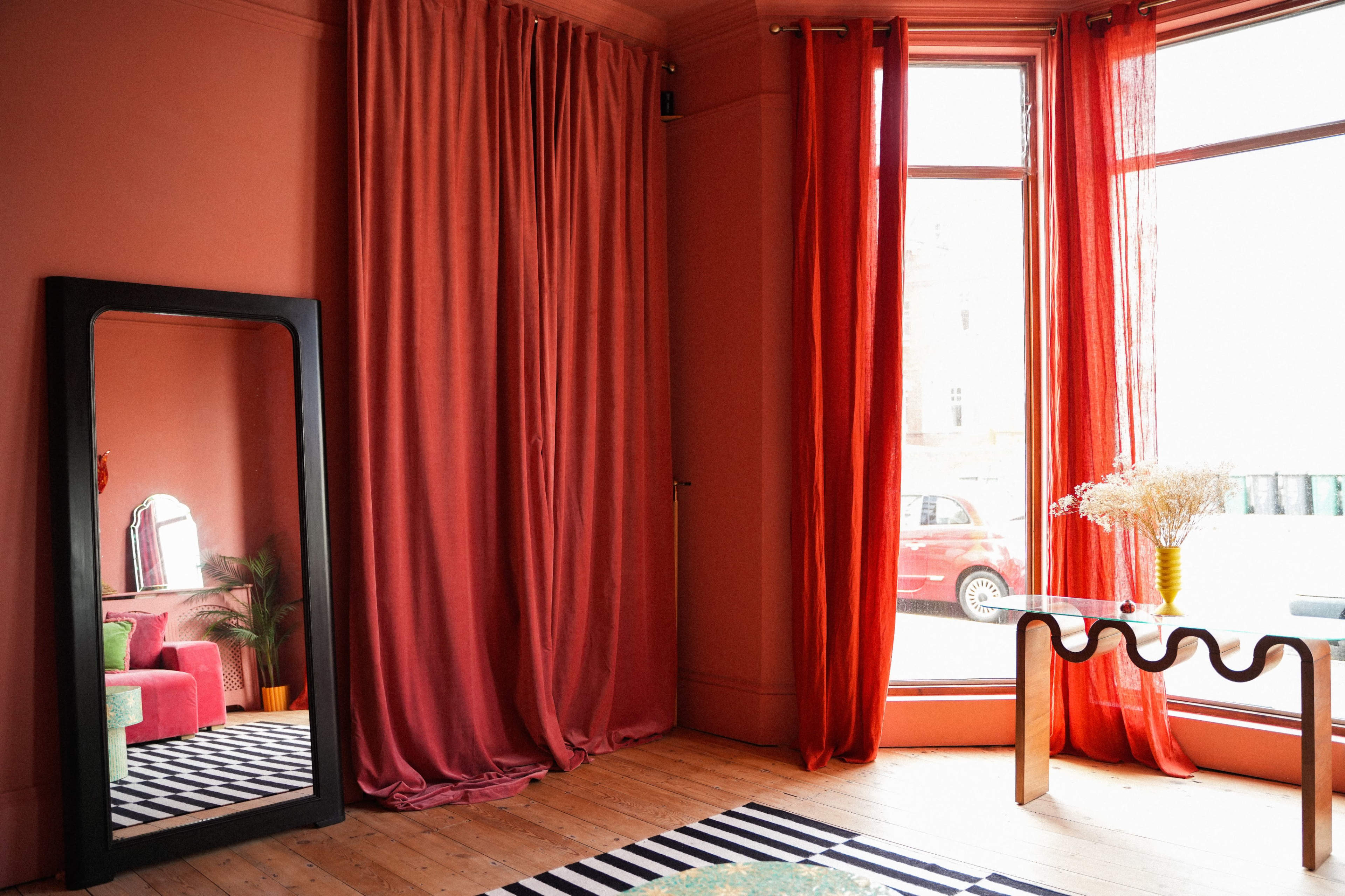 A spacious room features a large mirror, vibrant red curtains, a striped rug, and a small table by a window overlooking a parked car.