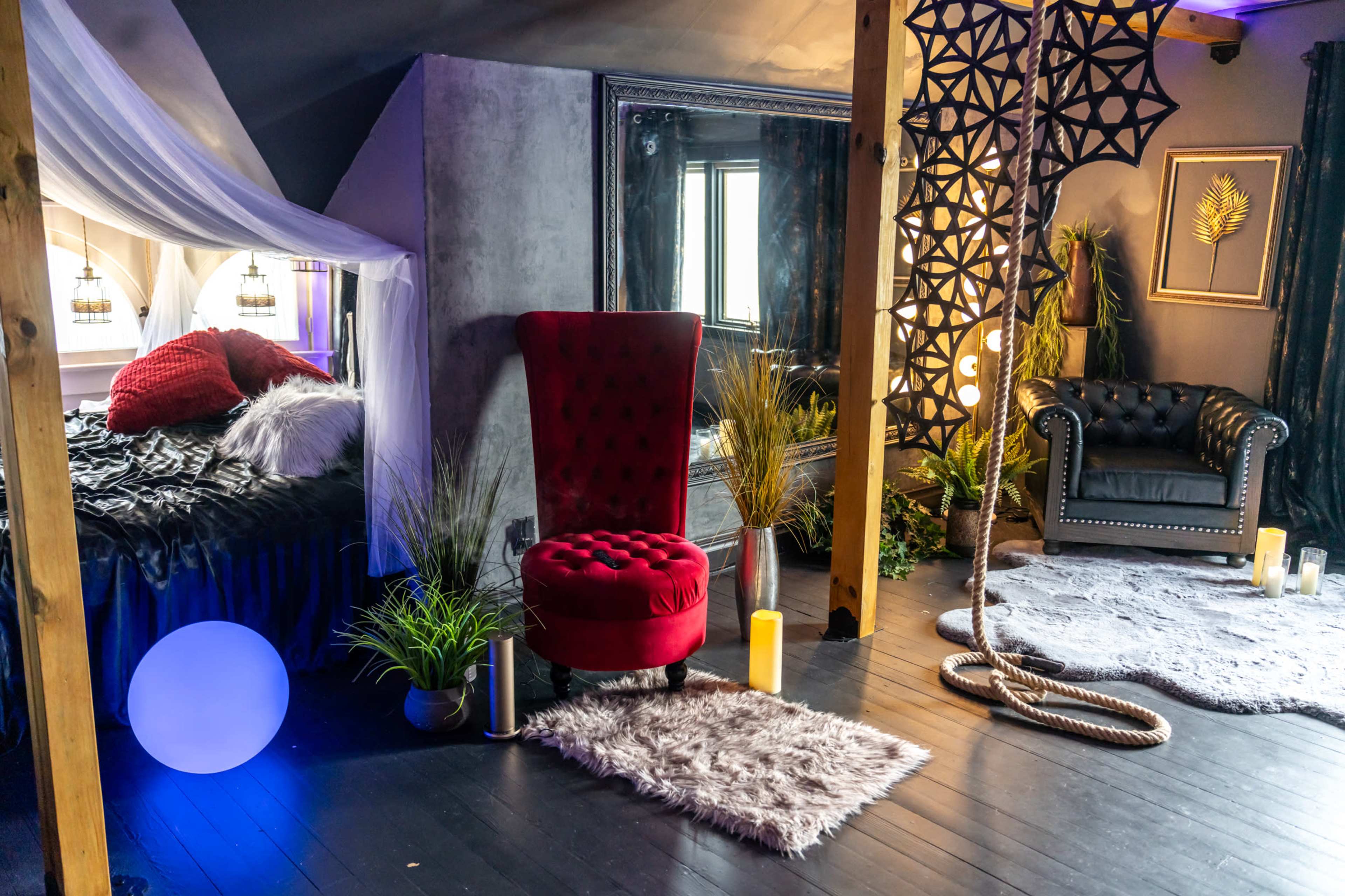 The room features a red velvet armchair, a black leather sofa, a bed with draped fabric, and decorative plants, all arranged on dark wooden flooring.