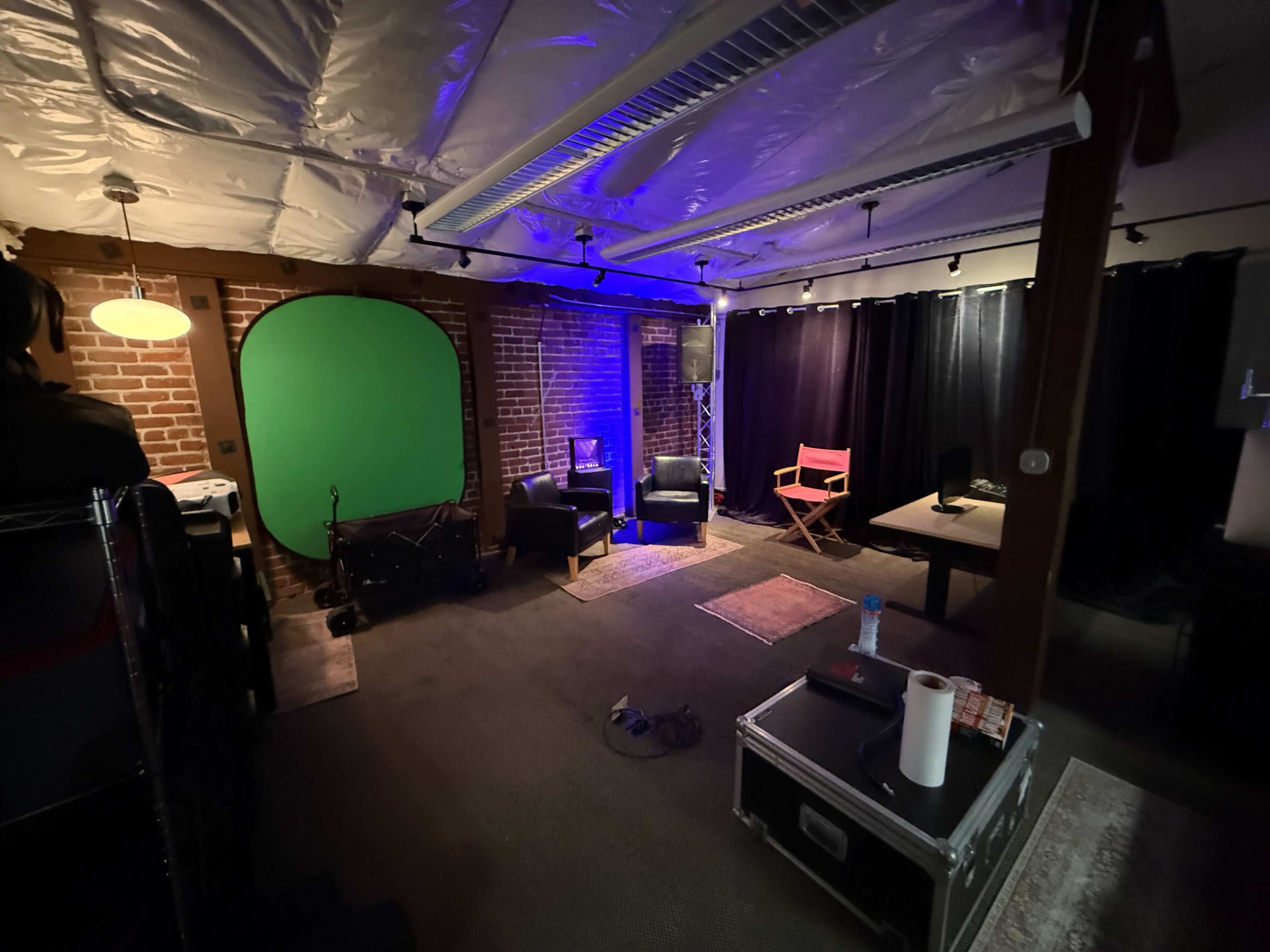 The image shows a studio space with exposed brick walls, green screen setup, two black chairs, a director's chair, and various equipment in a dimly lit environment.