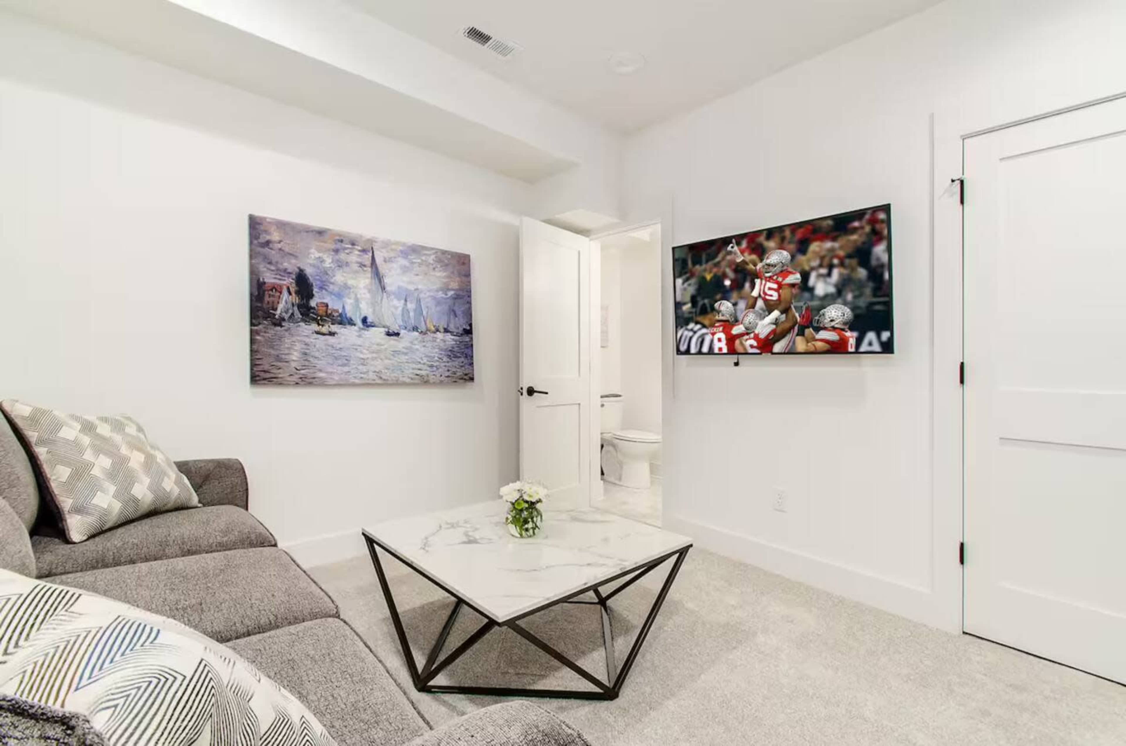 The image shows a compact living room featuring a gray sofa, a marble-topped coffee table, a television mounted on the wall, and a painting beside an open door leading to a bathroom.