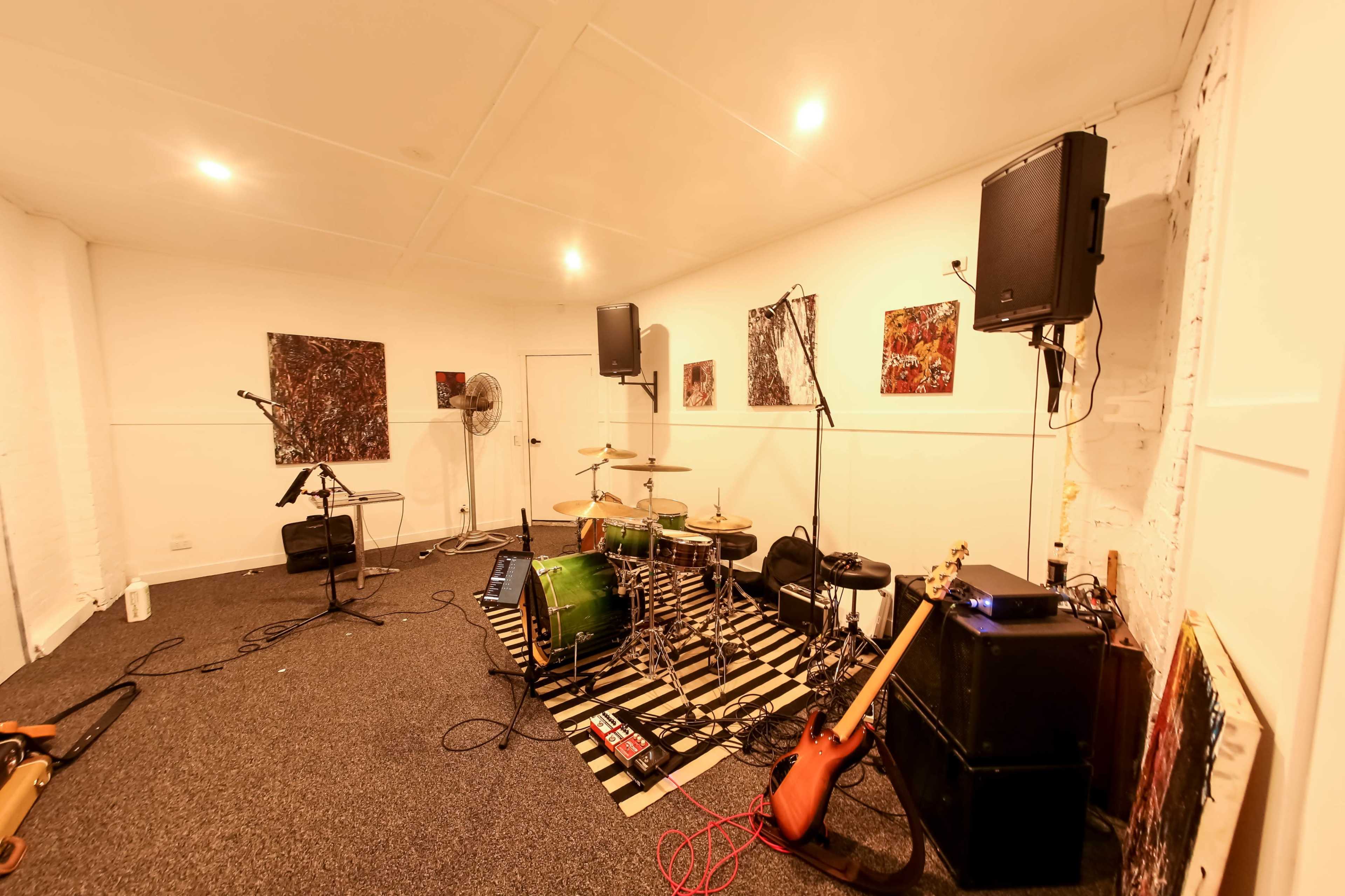 The image shows a music rehearsal space with a drum set, electric guitar, amplifiers, and sound equipment arranged along the walls.