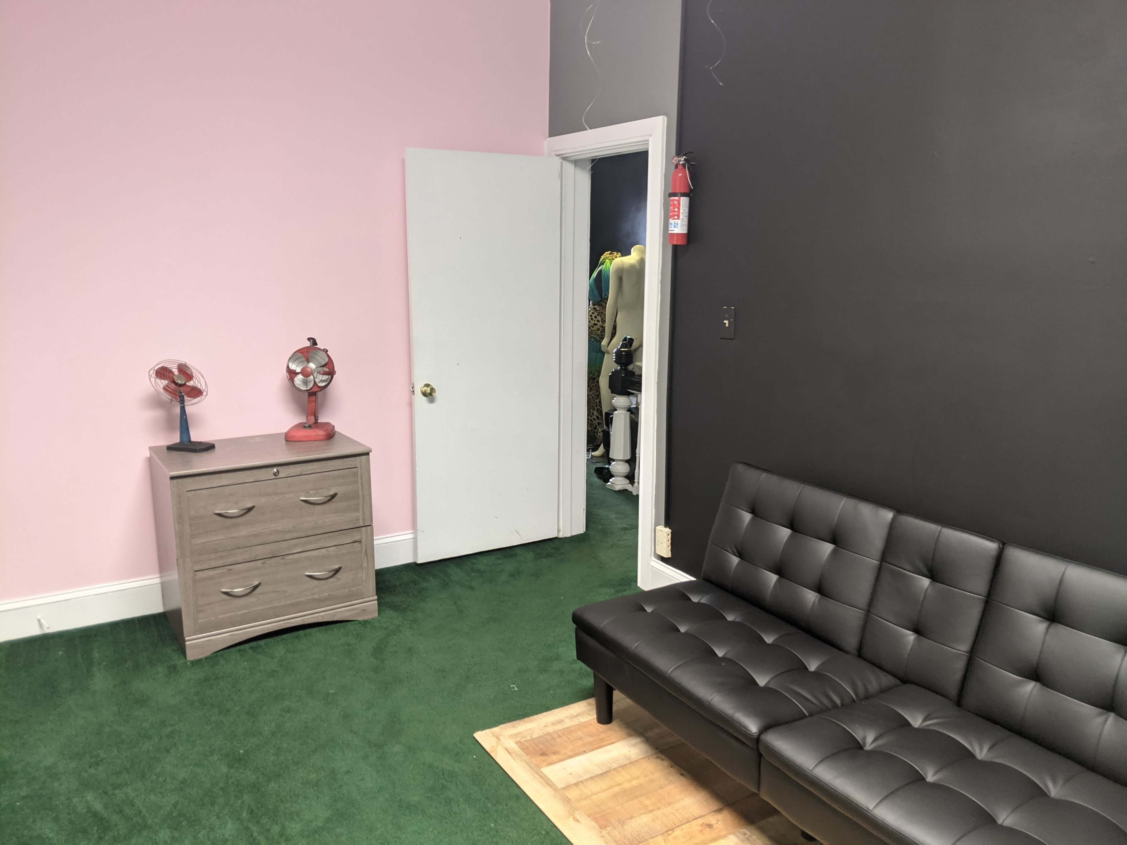 A room with a pink wall, a black wall, a gray dresser, a black couch, and a door leading to another area.