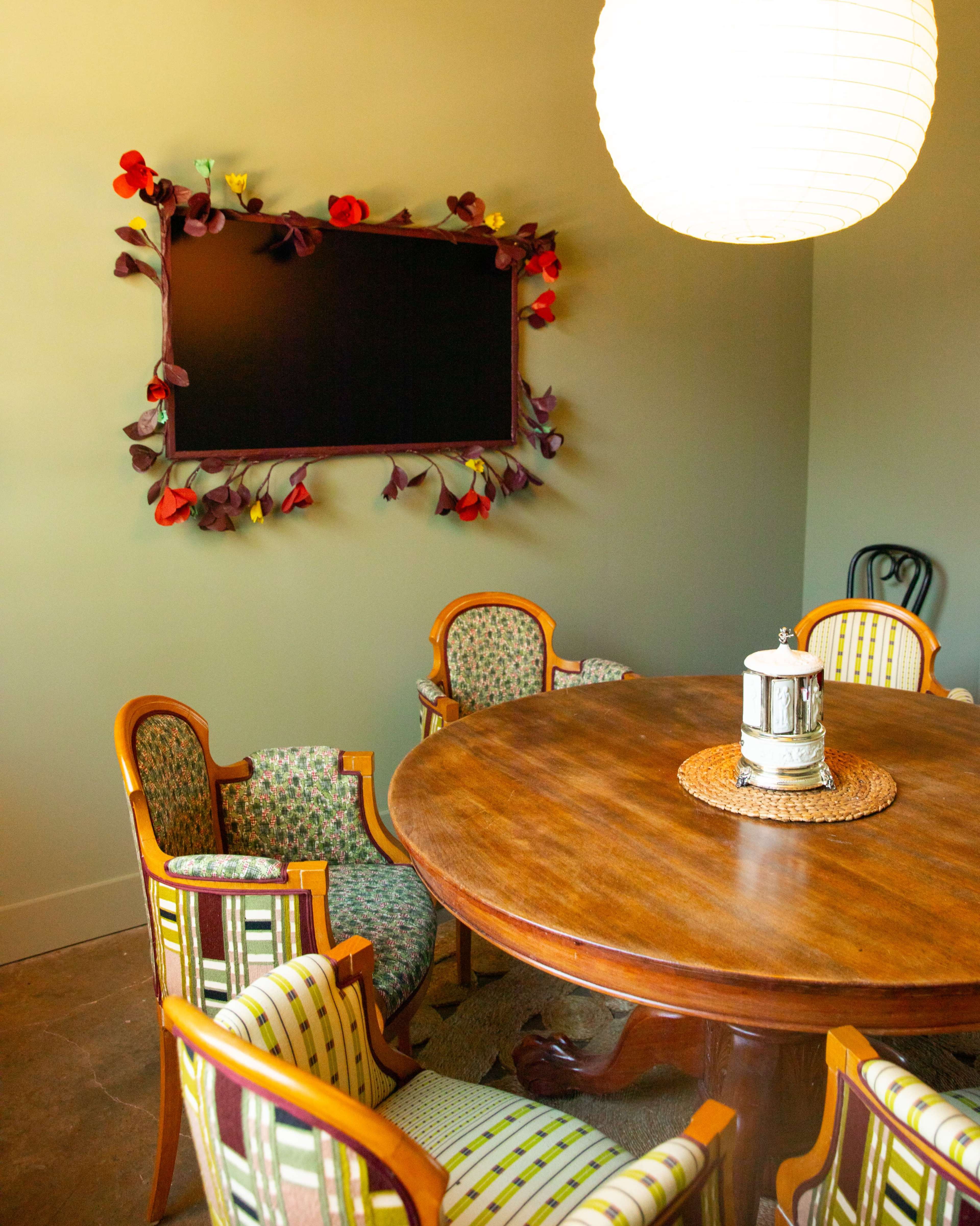 A round wooden dining table surrounded by patterned chairs sits beneath a large paper lantern and a floral-framed blackboard on the wall.