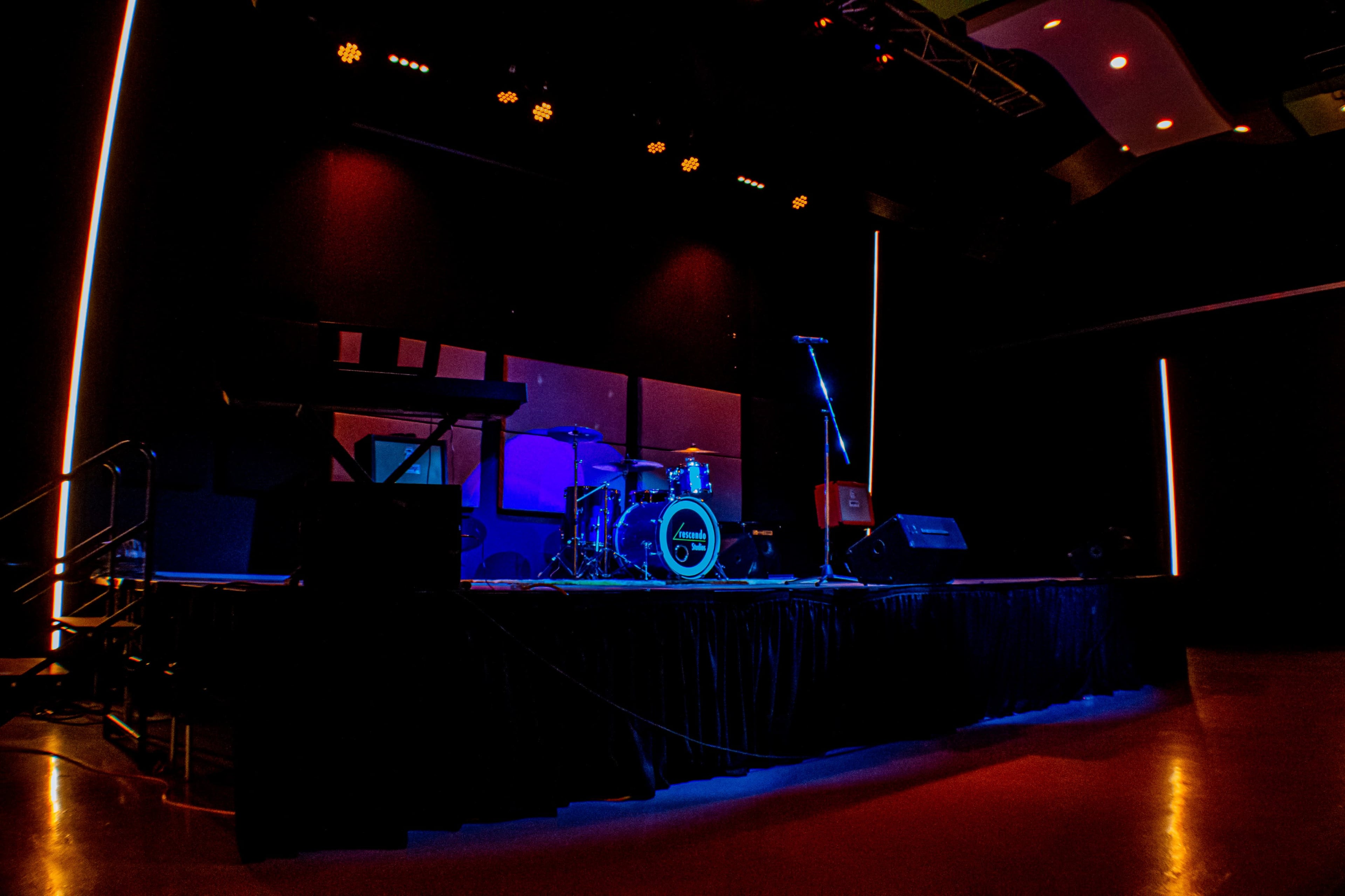 The stage is set for a musical performance, featuring a drum kit, microphone stand, and amplifiers, illuminated by colored lights.