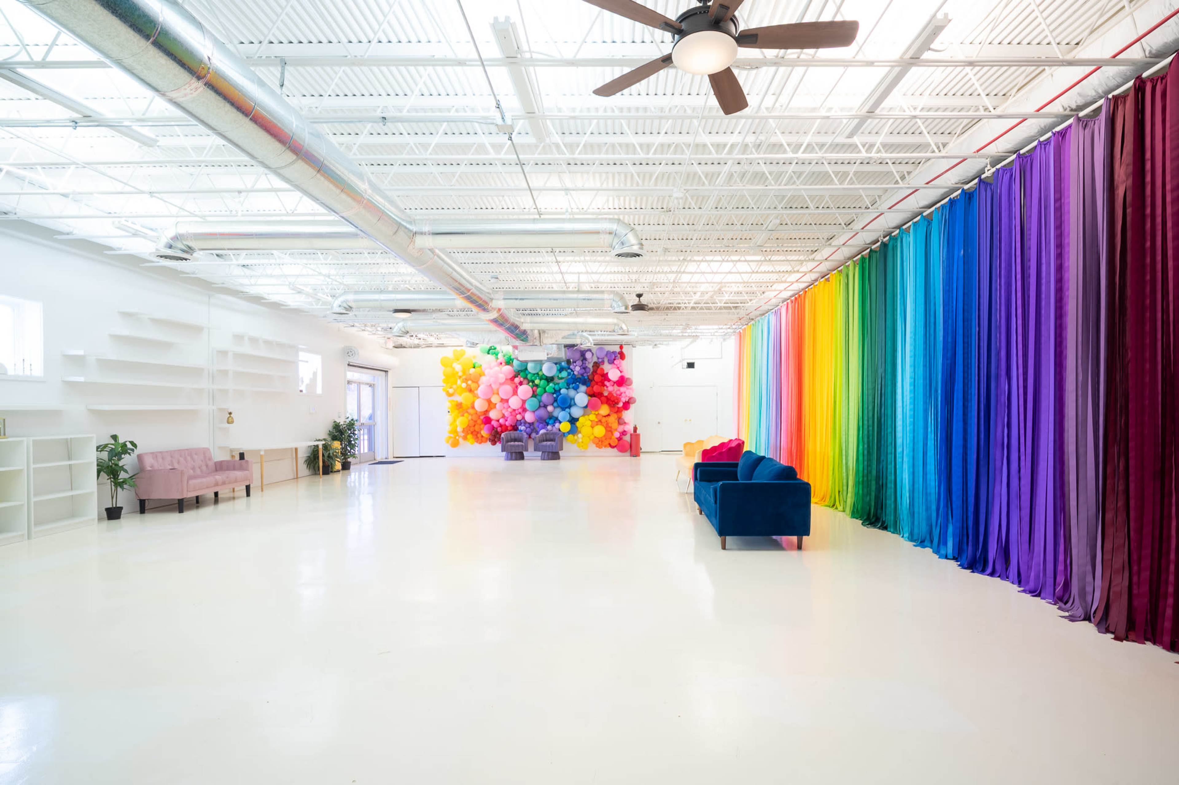 The spacious interior features a colorful balloon installation at one end and a curtain of vibrant fabrics on the other, with two blue sofas and a light-colored floor.