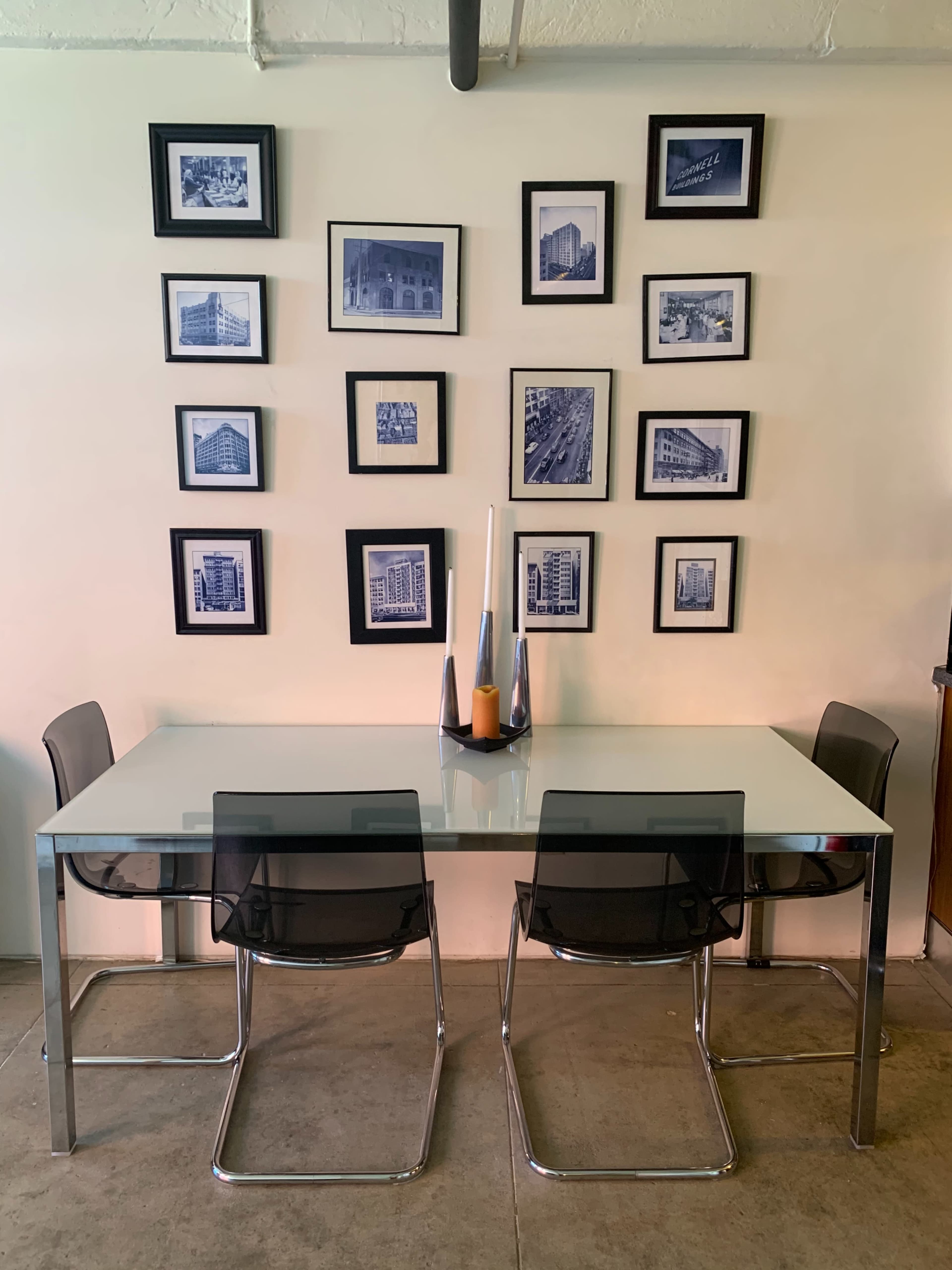 A modern dining table with four transparent chairs is positioned on a floor, beneath a wall featuring a grid of framed black-and-white photographs.