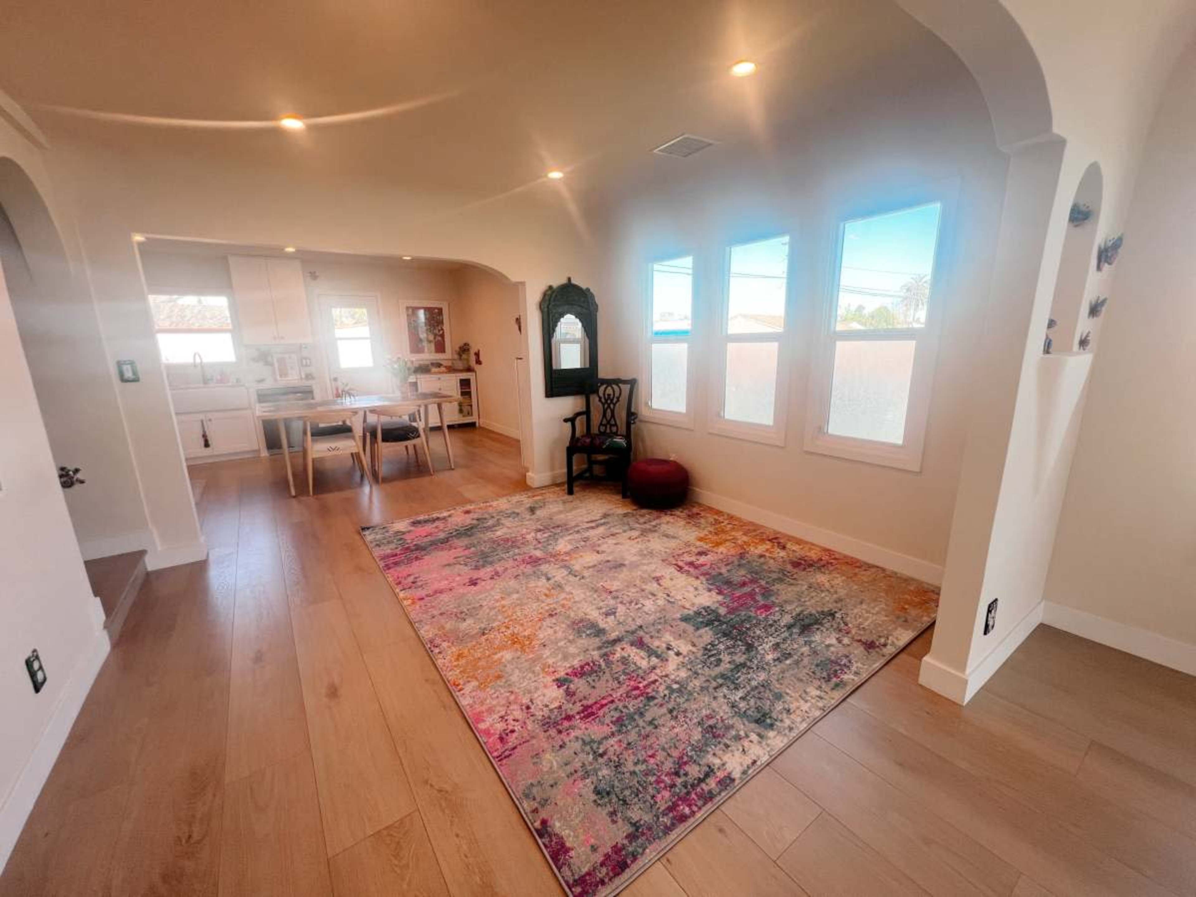 The image shows a bright, minimally furnished living space featuring large windows, a vibrant area rug, and a wooden dining table in the adjacent kitchen area.
