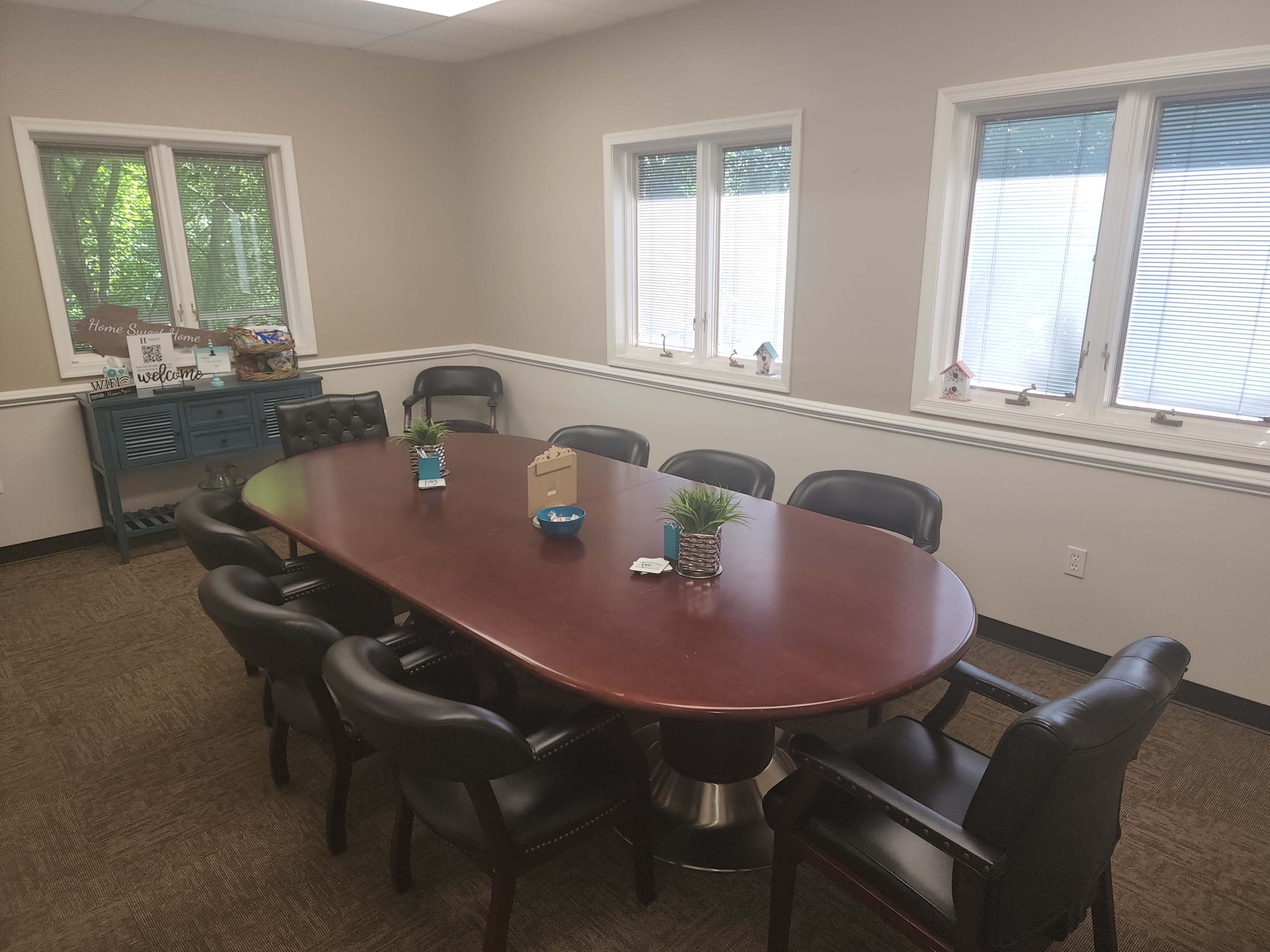 A conference room with a large oval wooden table surrounded by black leather chairs, and windows letting in natural light.