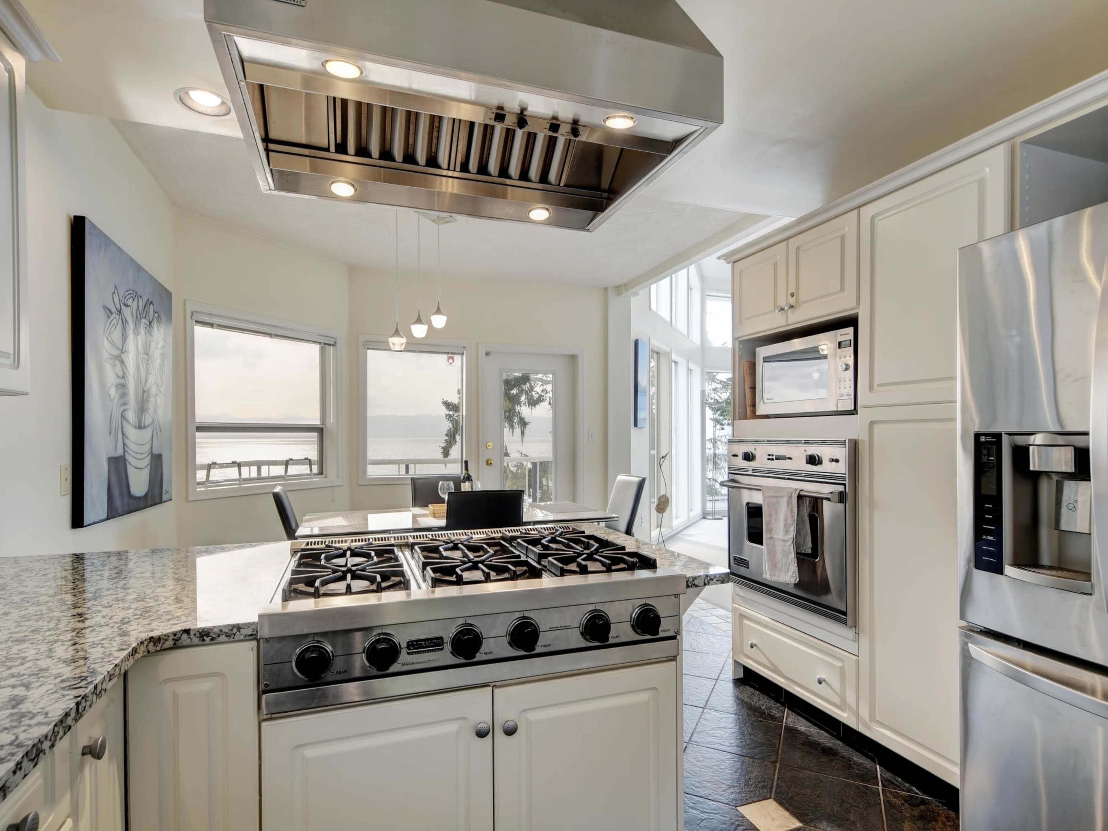 A modern kitchen features stainless steel appliances, a large gas range, and a view of a waterfront through tall windows.