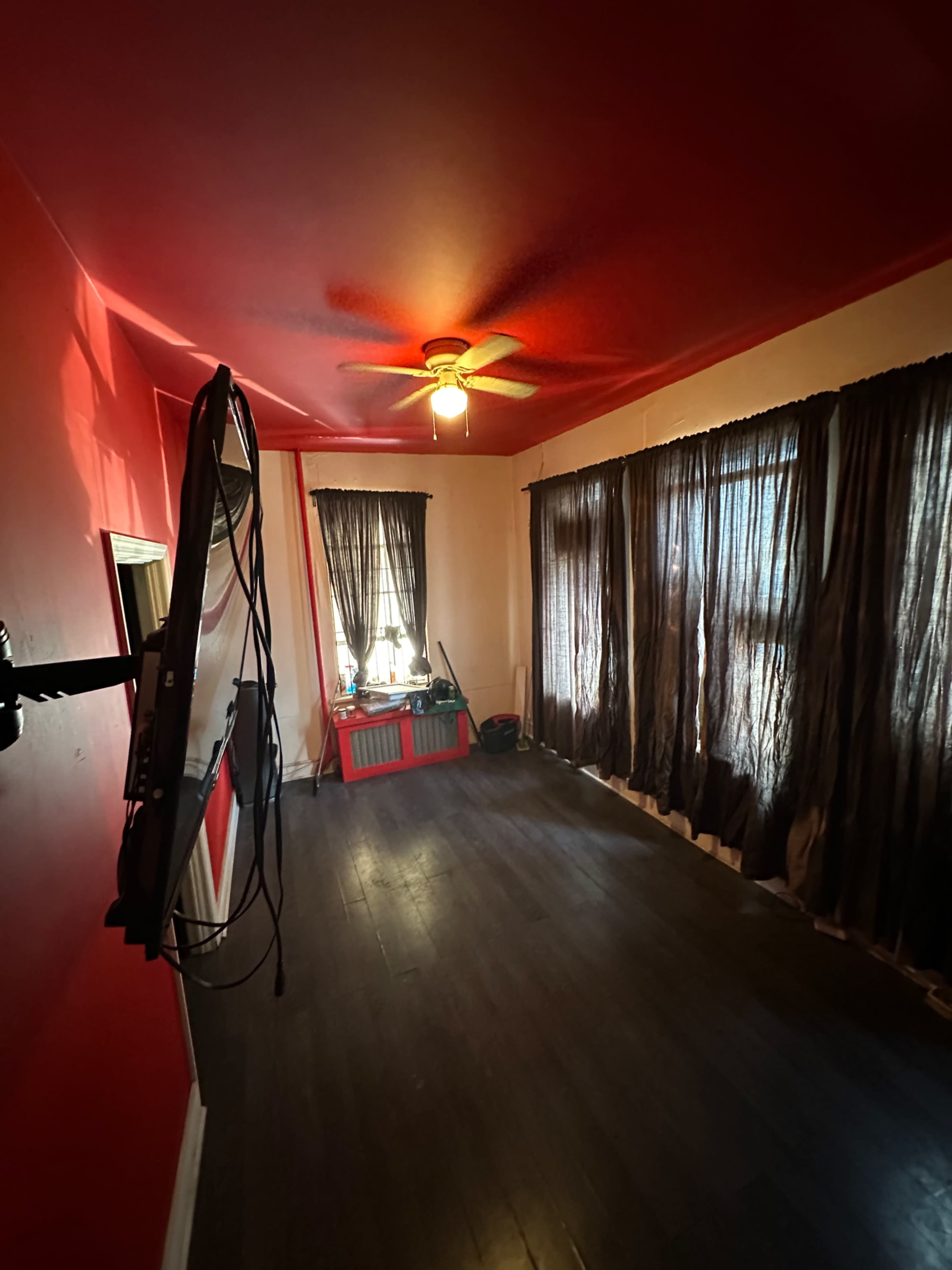 A room with red walls, a ceiling fan, a mounted television, and long dark curtains over windows.
