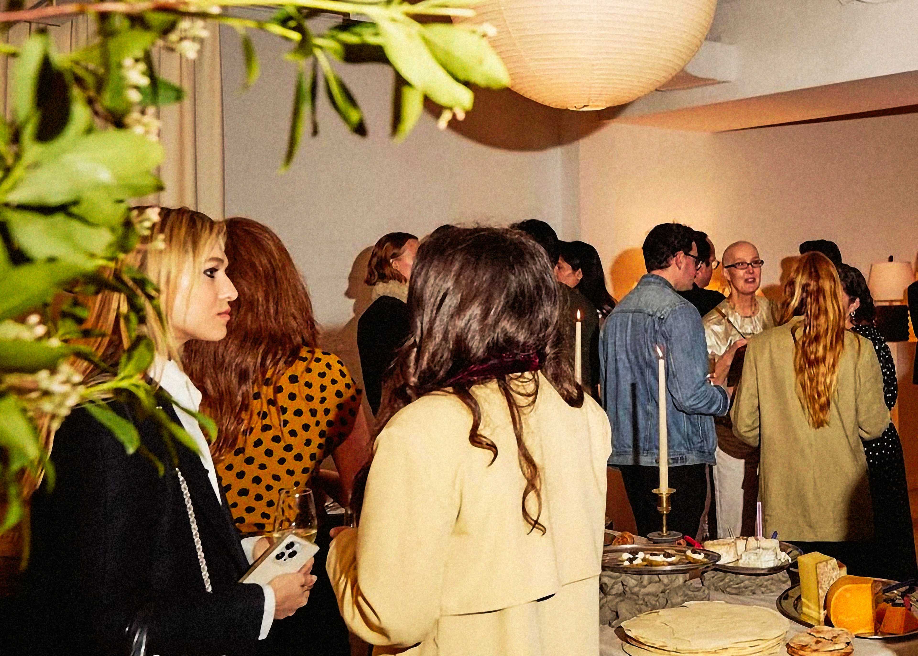 A gathering of people in a room with a table of food and drinks, surrounded by soft lighting and decorative plants.