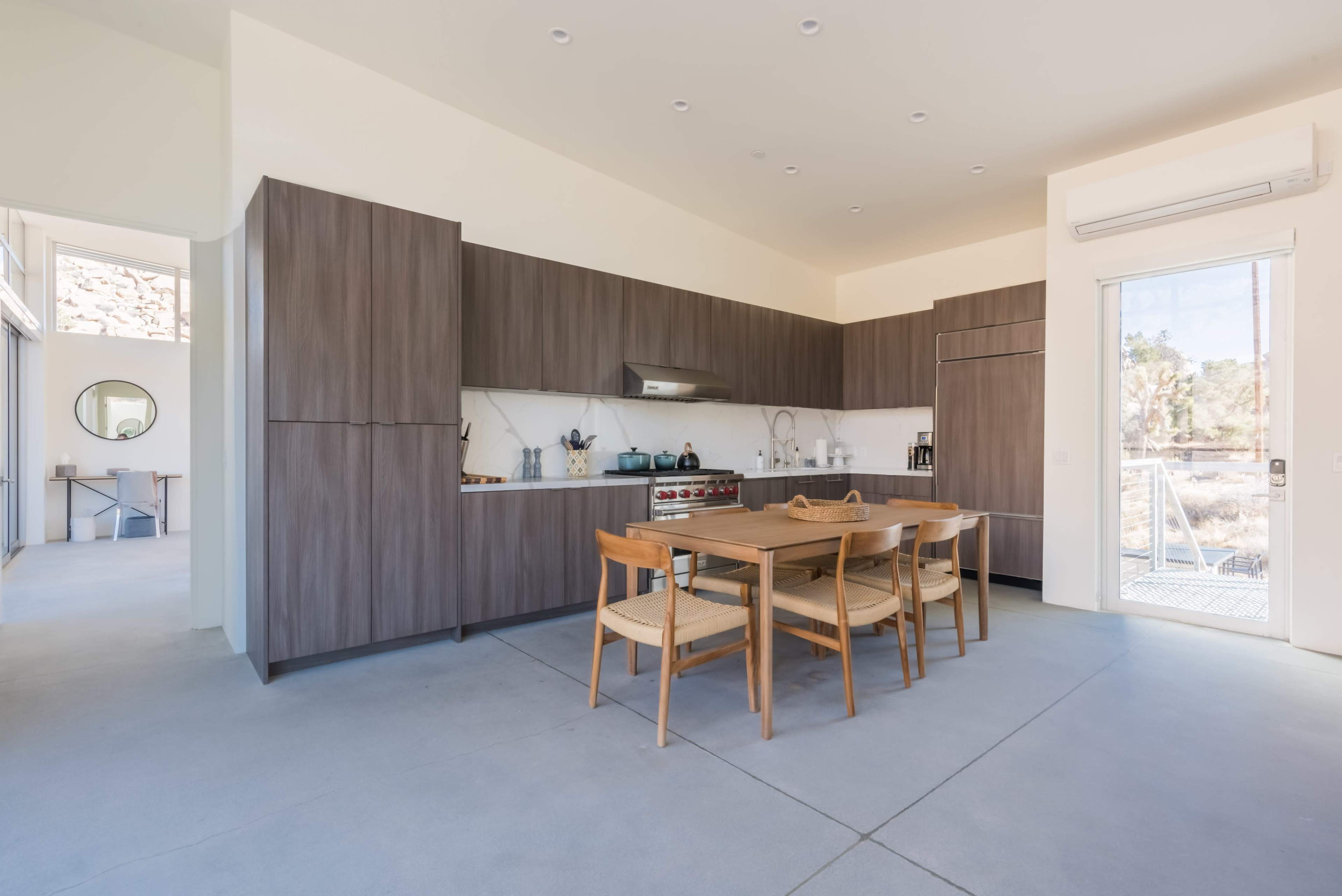 The image shows a modern kitchen with a large wooden dining table and minimalist cabinetry, featuring a linear layout and natural light coming from a nearby door.