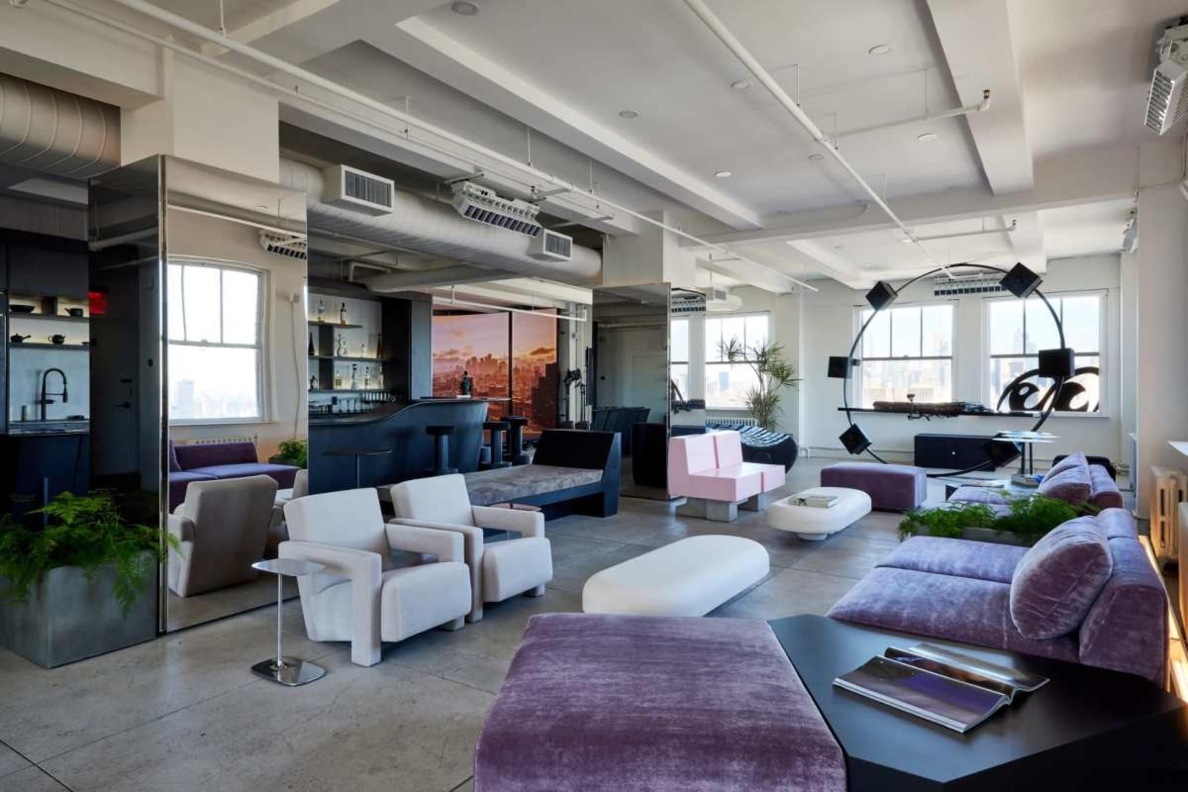 The image shows a modern living space with various seating arrangements, large windows, and an open layout that includes a kitchenette area.