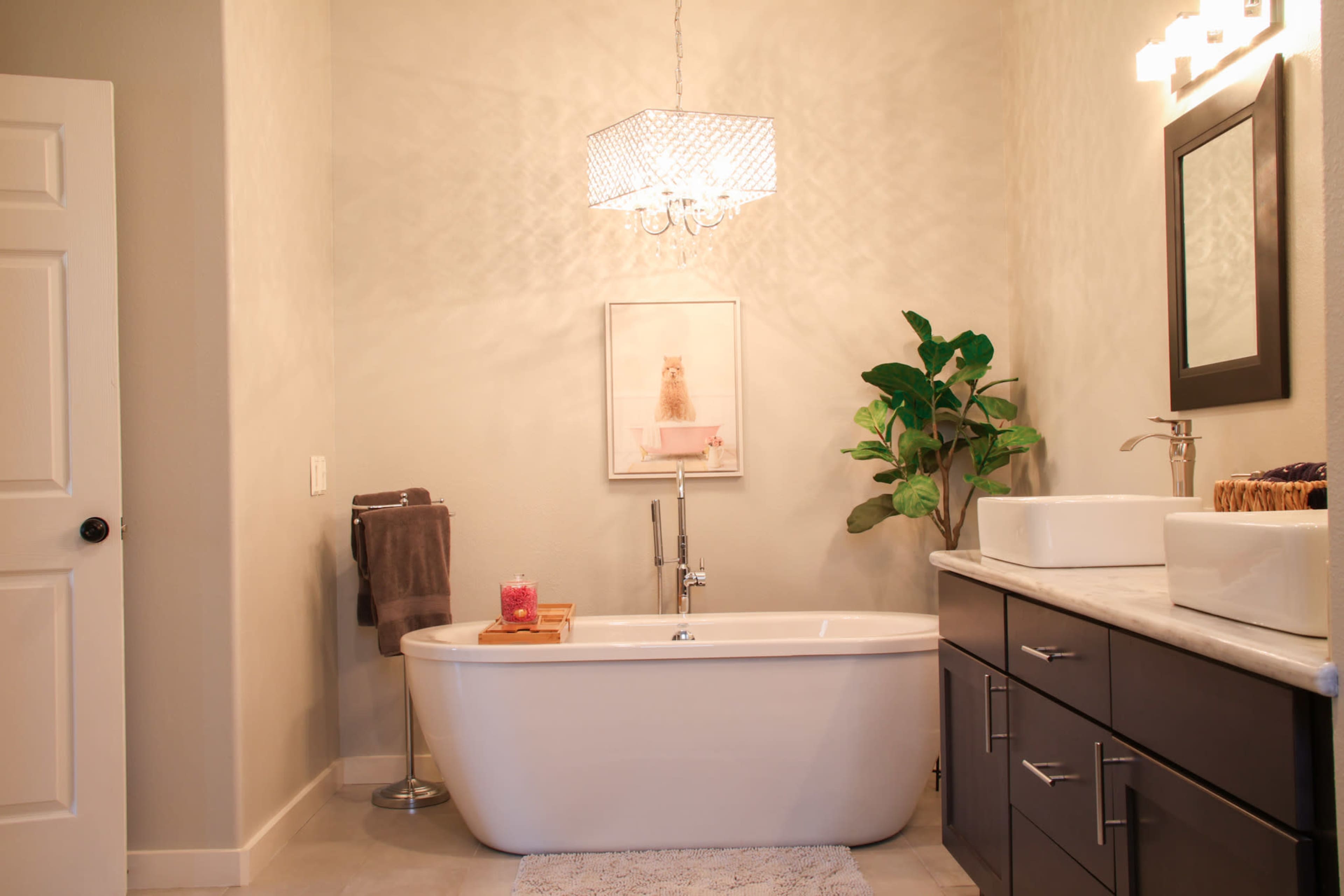 A modern bathroom features a freestanding tub, a chandelier, a plant, and dual sinks with dark cabinetry.
