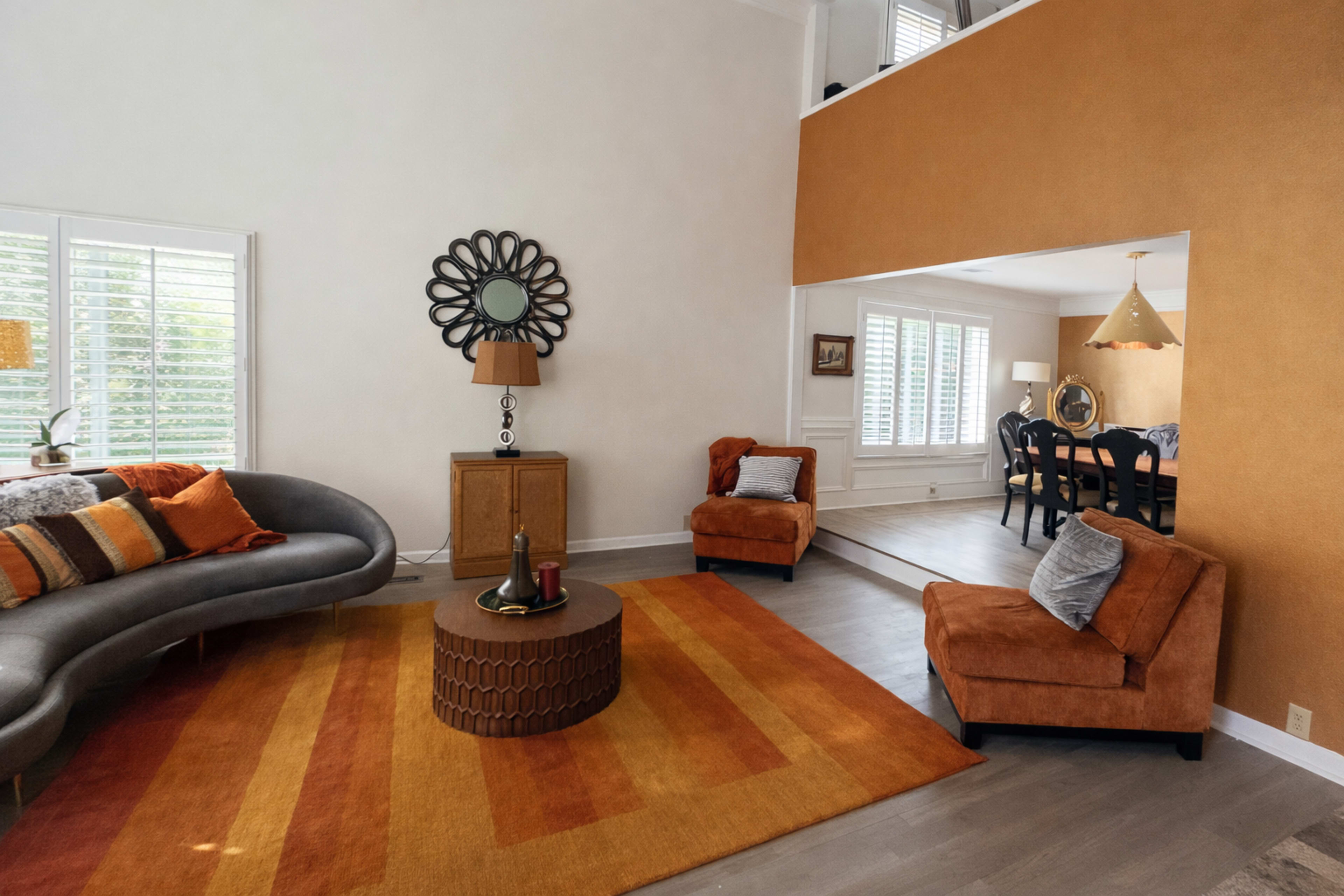 The image shows a modern living room with a curved gray sofa, orange armchair, a round coffee table, and a striped area rug, along with a dining area visible in the background.