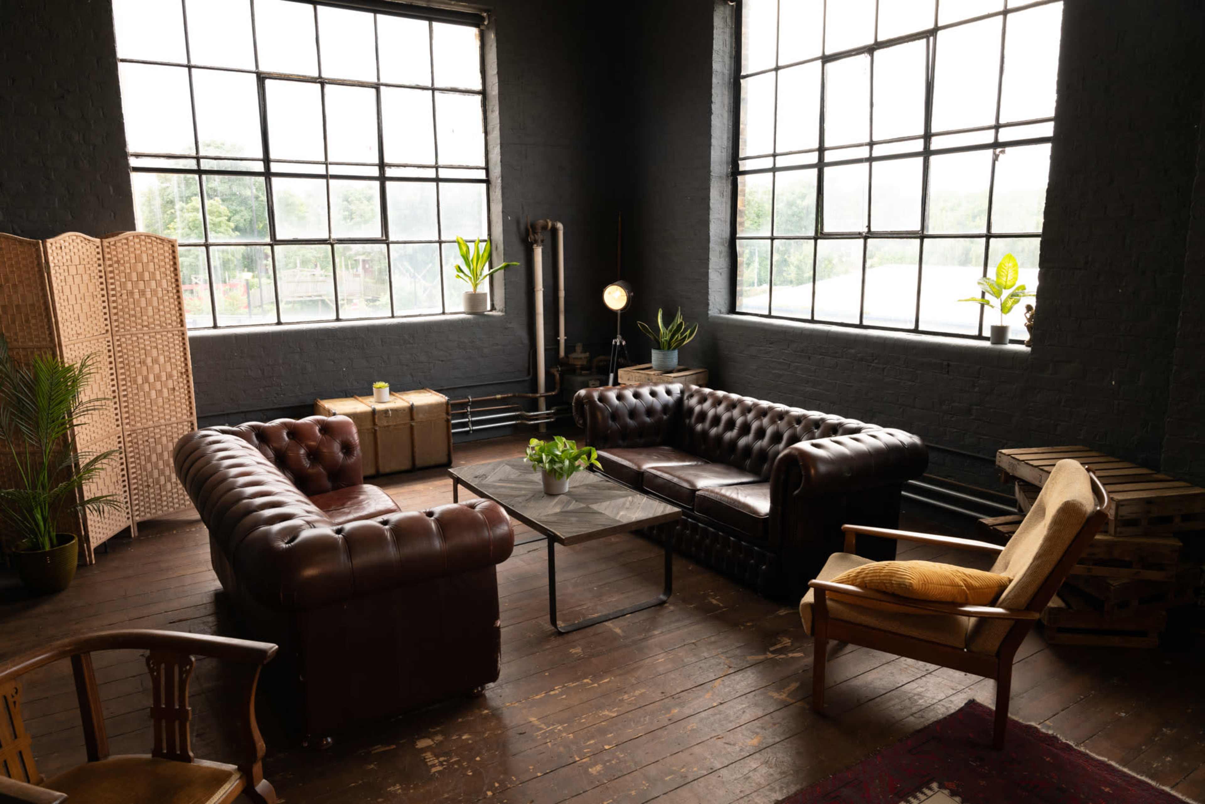 A spacious room with large windows, featuring two brown leather sofas, a glass coffee table, and a mix of greenery and wooden furniture.