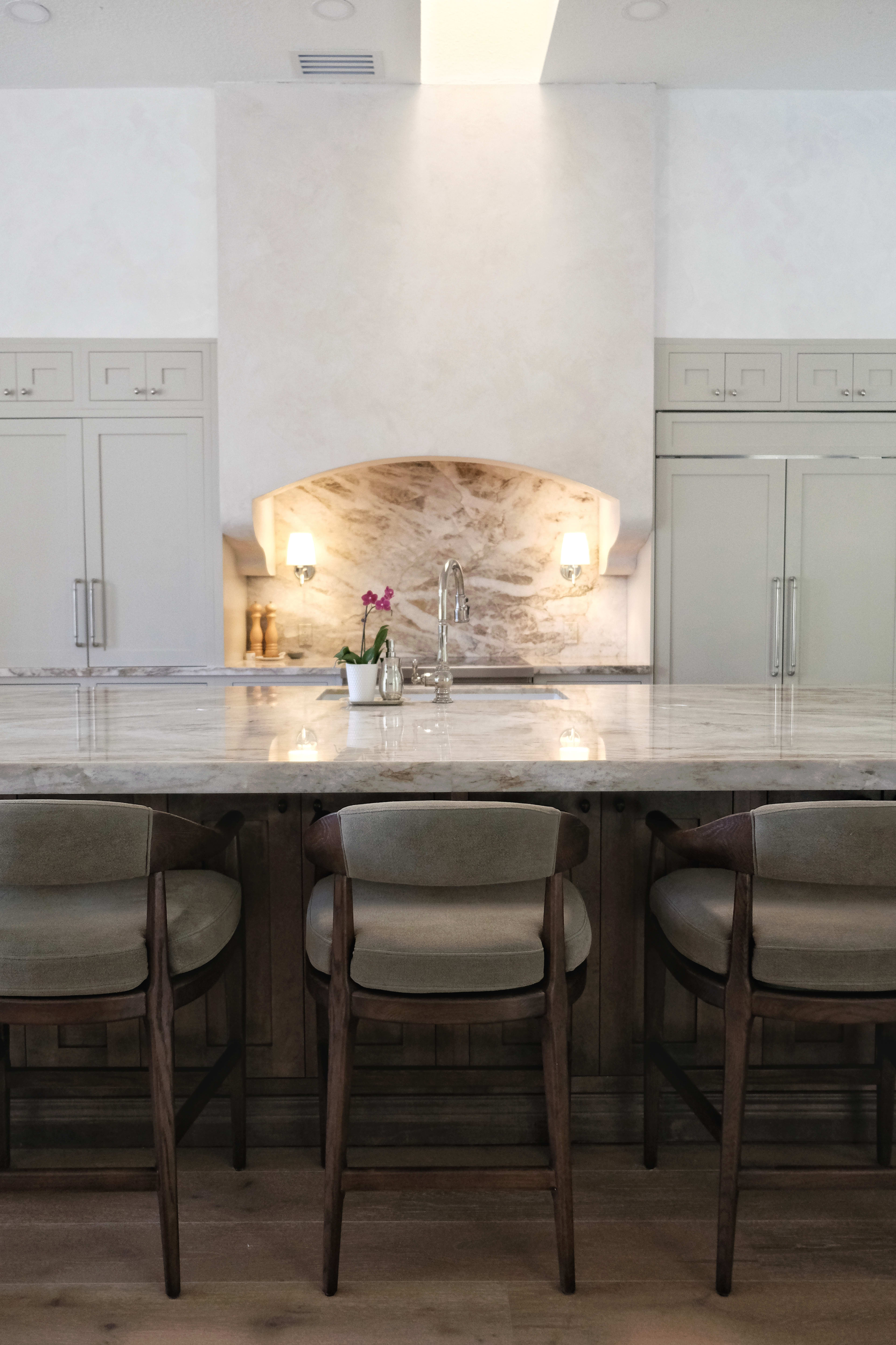 A modern kitchen features a large marble countertop with wooden bar stools and a stone backsplash behind a sink.