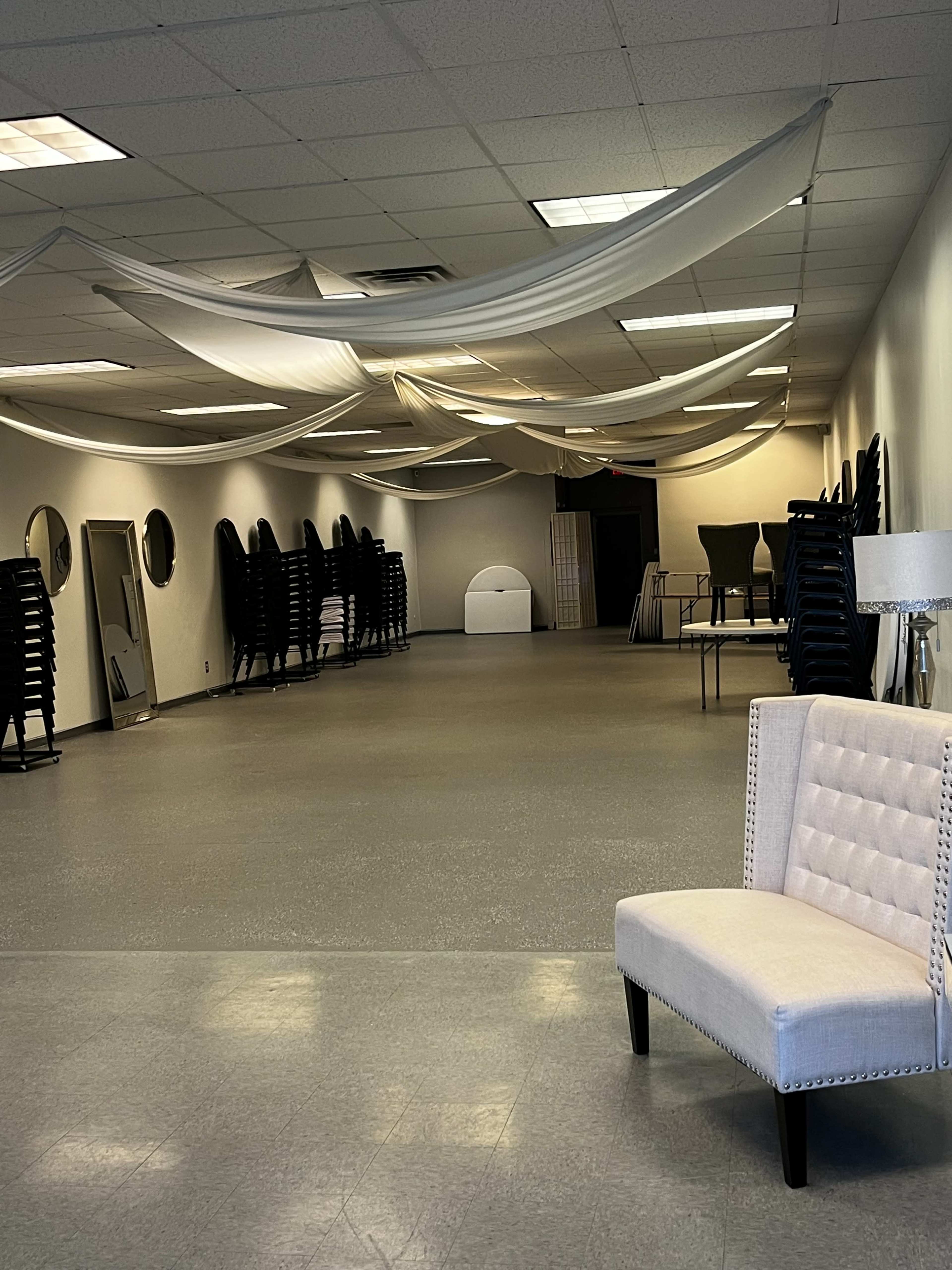 An empty event space with fabric draped from the ceiling, stacked chairs against the wall, and a decorative chair in the foreground.