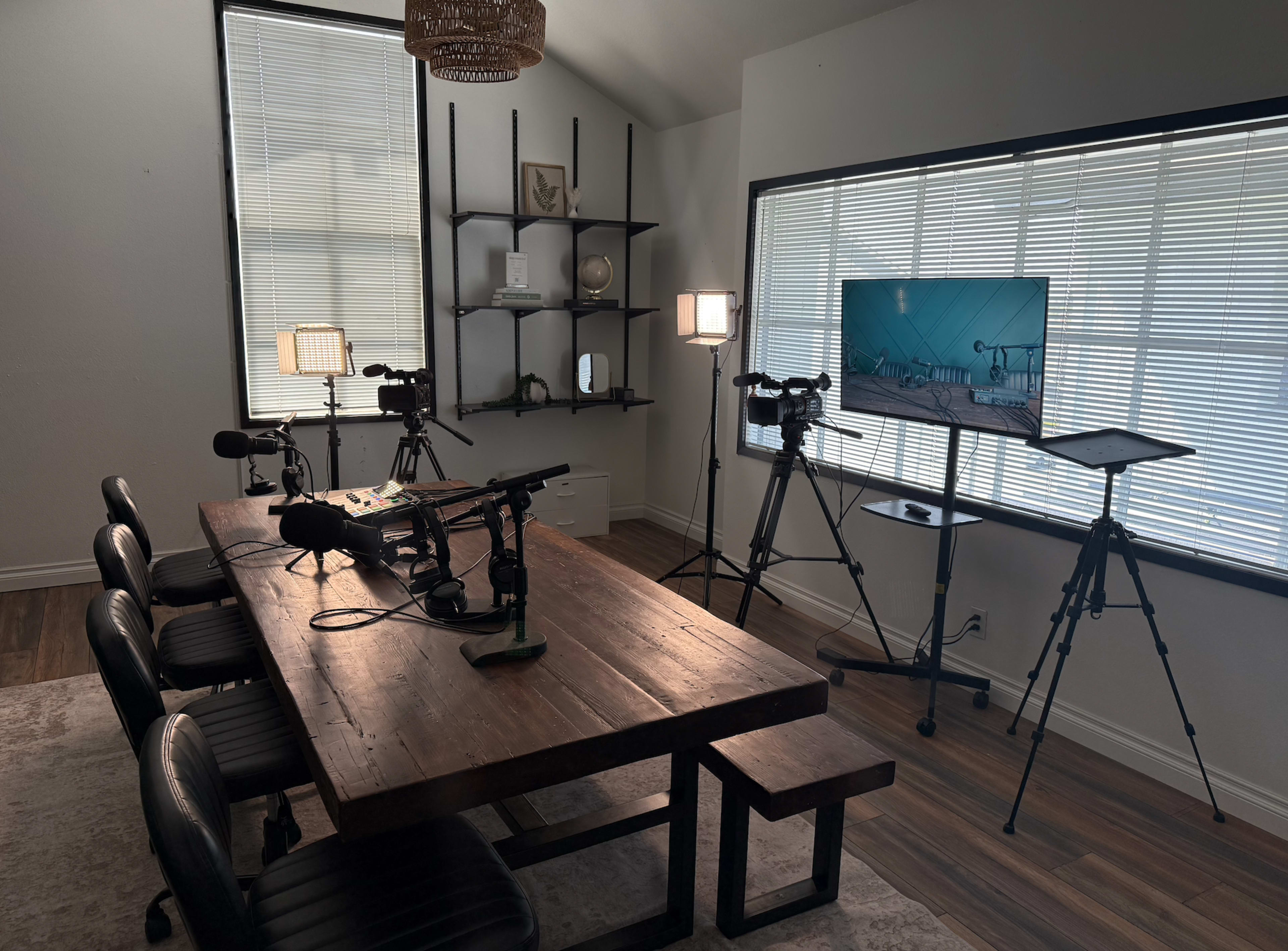 The image shows a well-lit recording studio with a large wooden table, microphones, cameras on tripods, and a monitor displaying a setup for video production.
