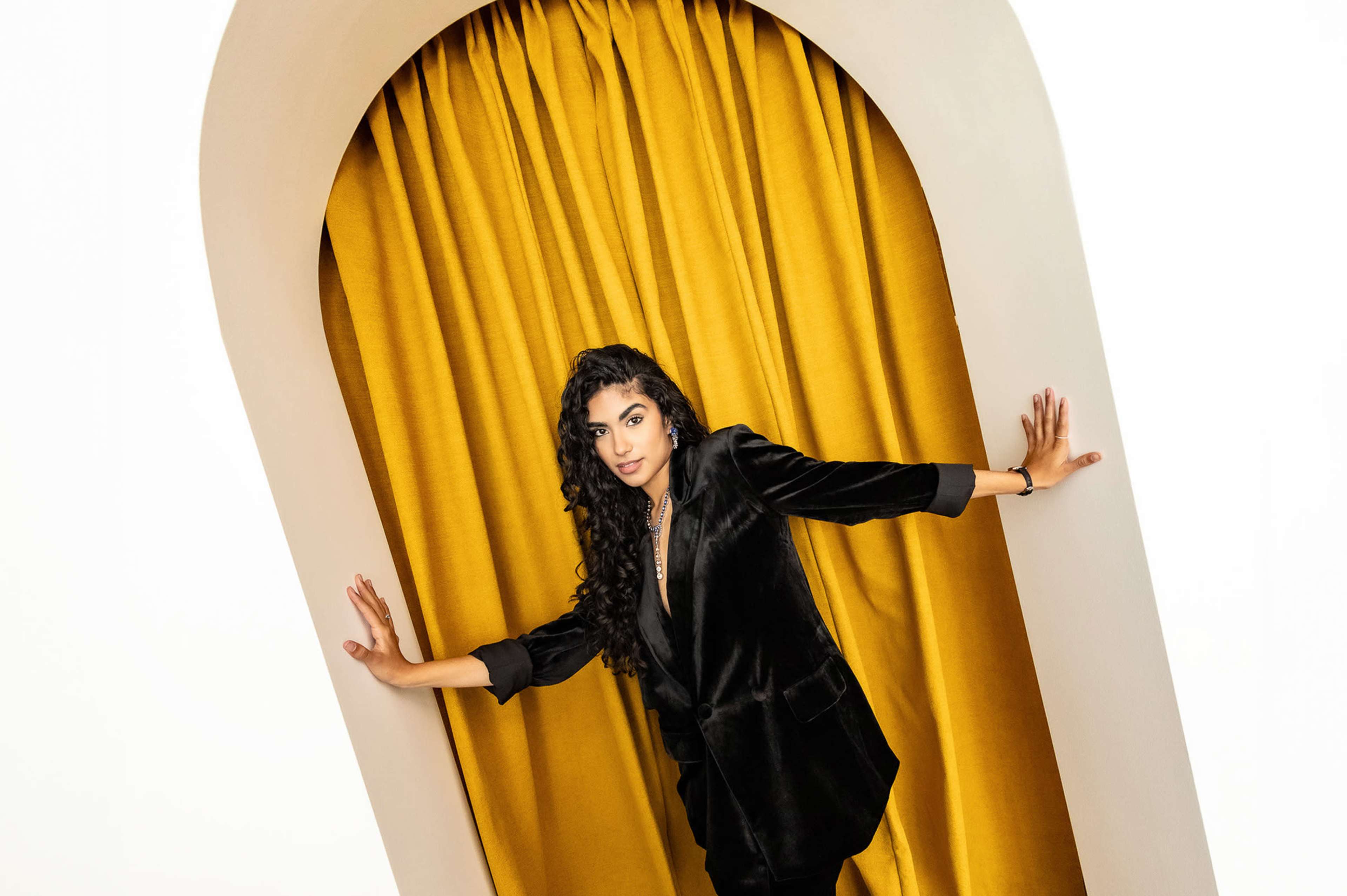 A woman with long, curly hair poses in front of a golden curtain framed by an arched doorway.