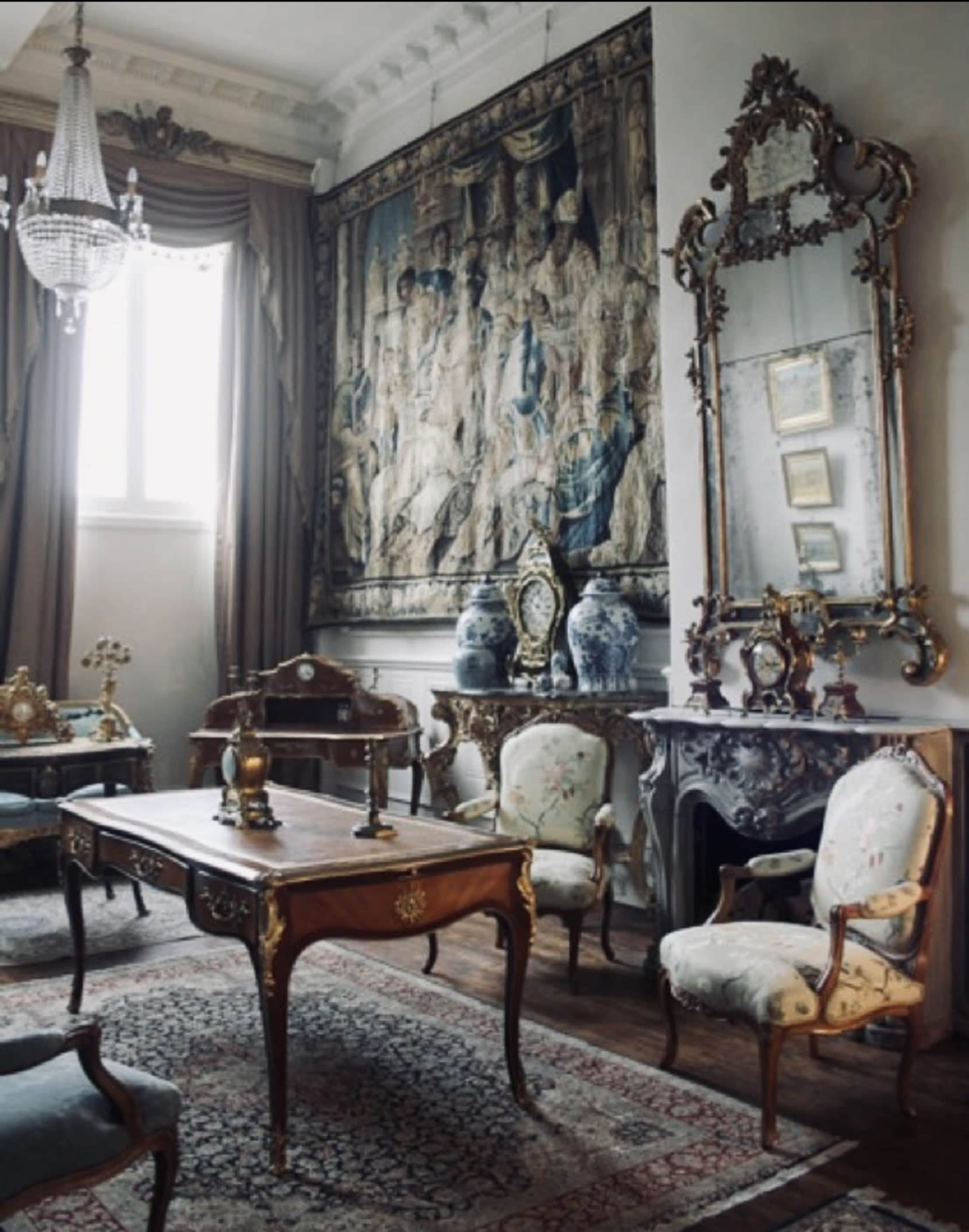 An elegantly furnished room featuring antique furniture, a large tapestry on the wall, and a chandelier hanging from the ceiling.
