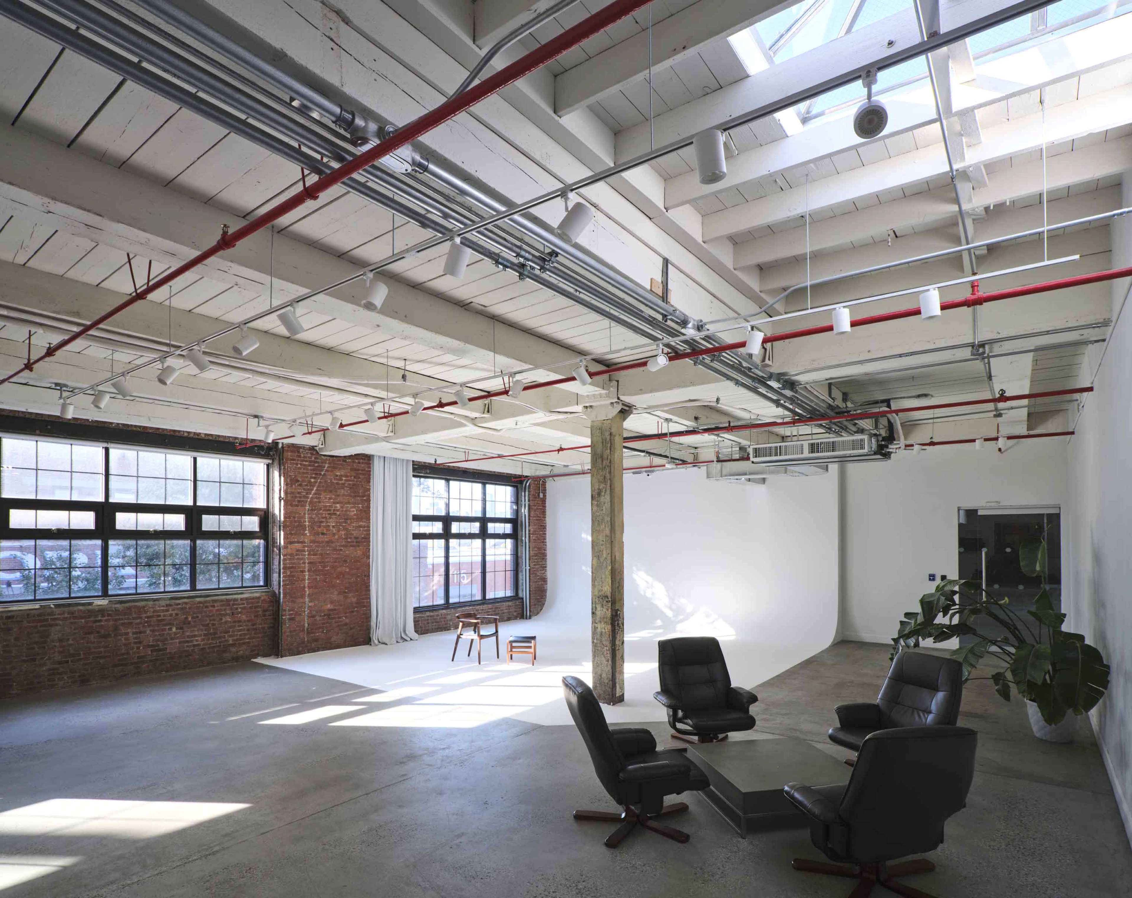 A spacious, loft-style interior with high ceilings, large windows, exposed brick walls, and a few pieces of furniture arranged in the center.