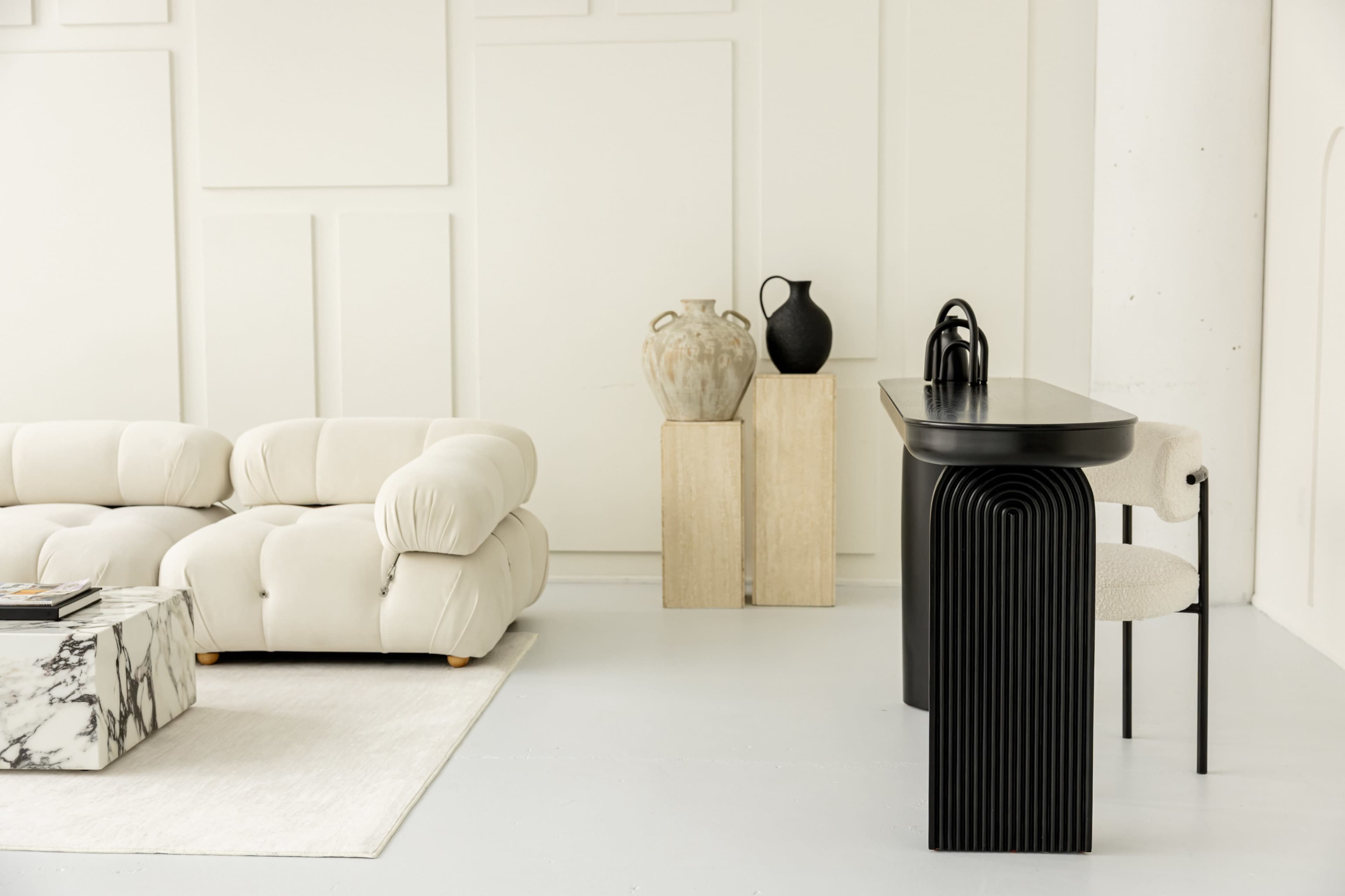 The image shows a minimalist interior space featuring a plush cream-colored sofa, a marble coffee table, and a sleek black bar with matching stools, alongside decorative vases on pedestals.