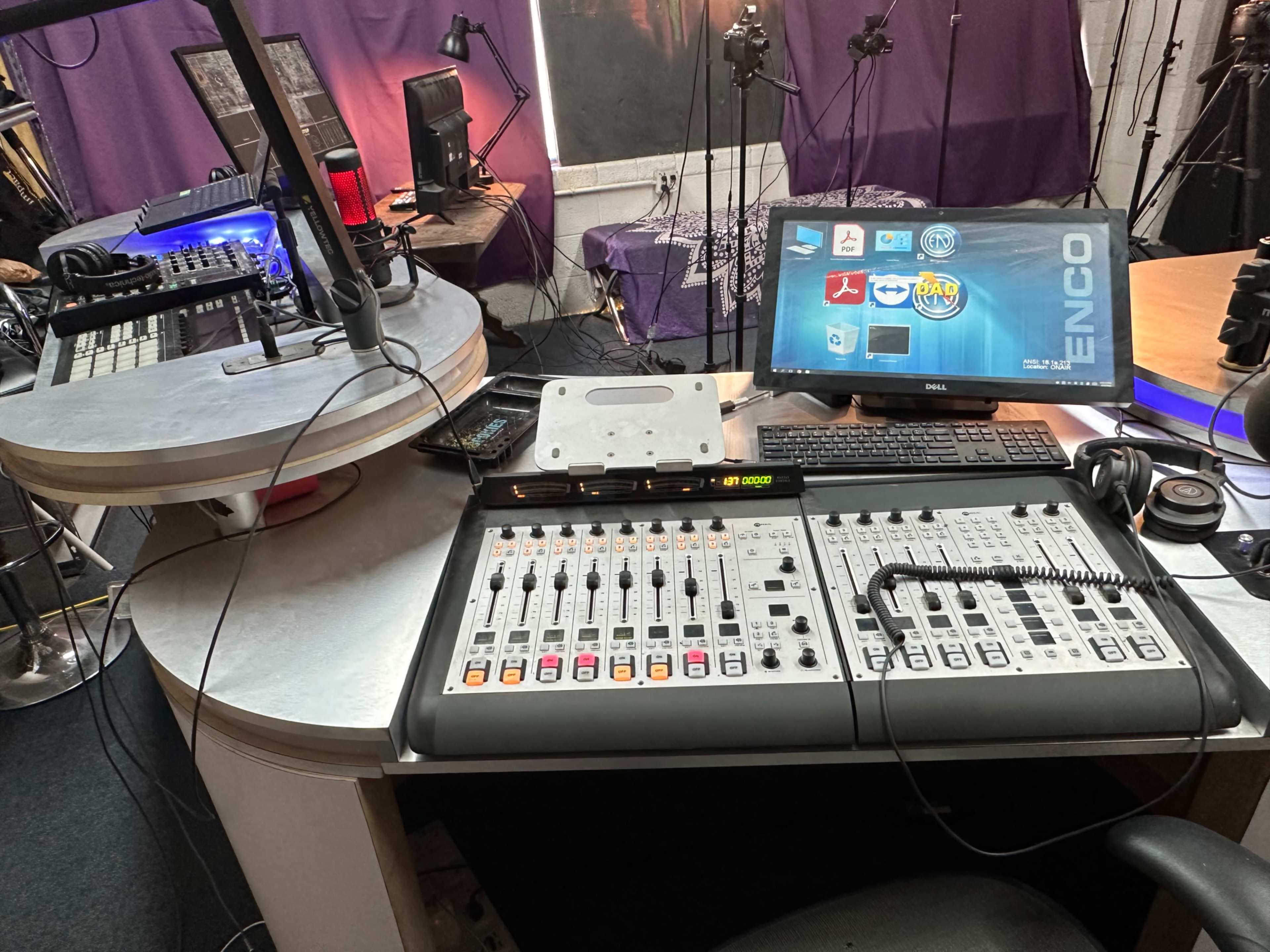 The image shows a radio studio setup with a mixing console, a computer monitor displaying software, and various audio equipment in a well-organized workspace.