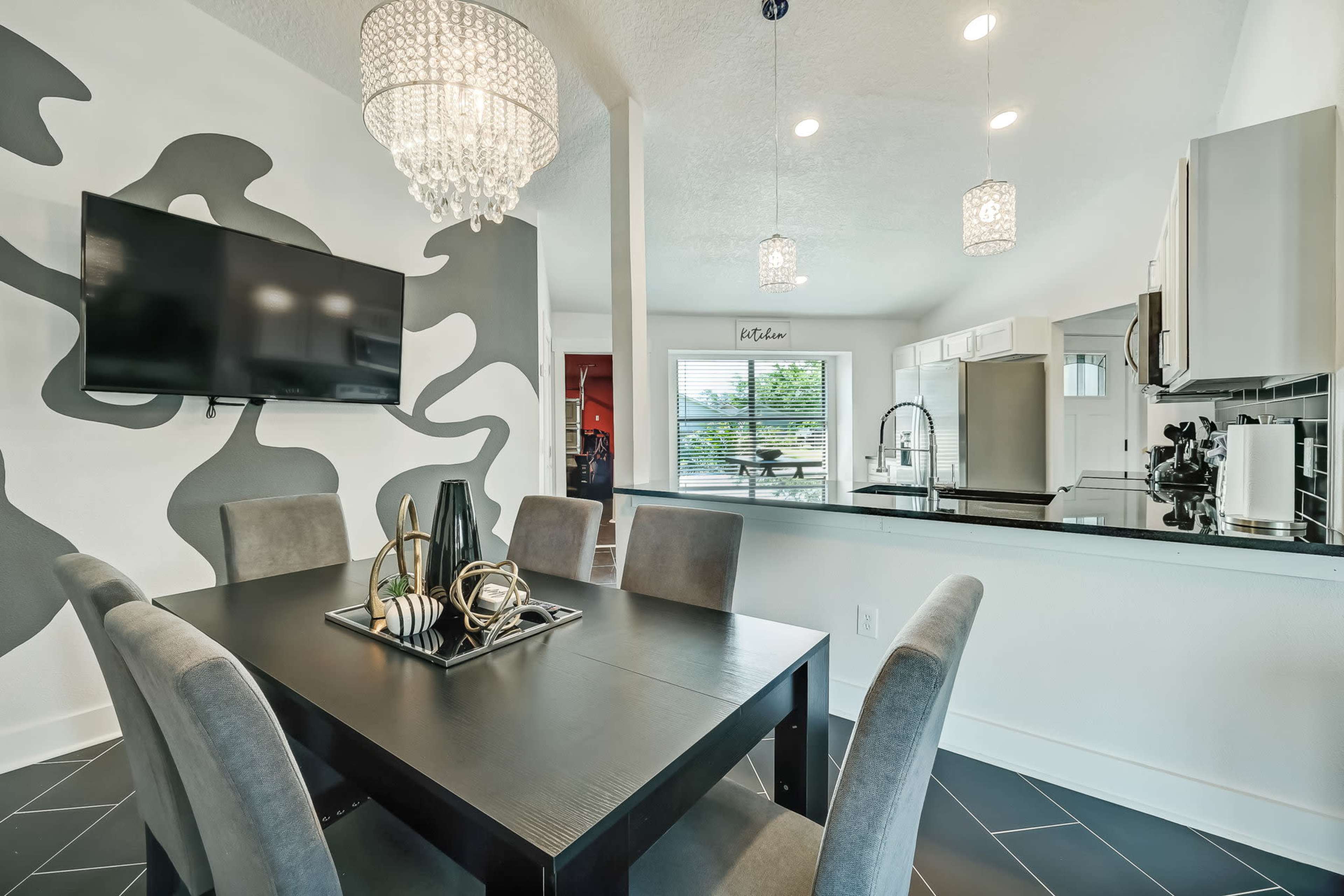 A dining area features a black table surrounded by gray chairs, with a large wall-mounted TV and a modern chandelier overhead.