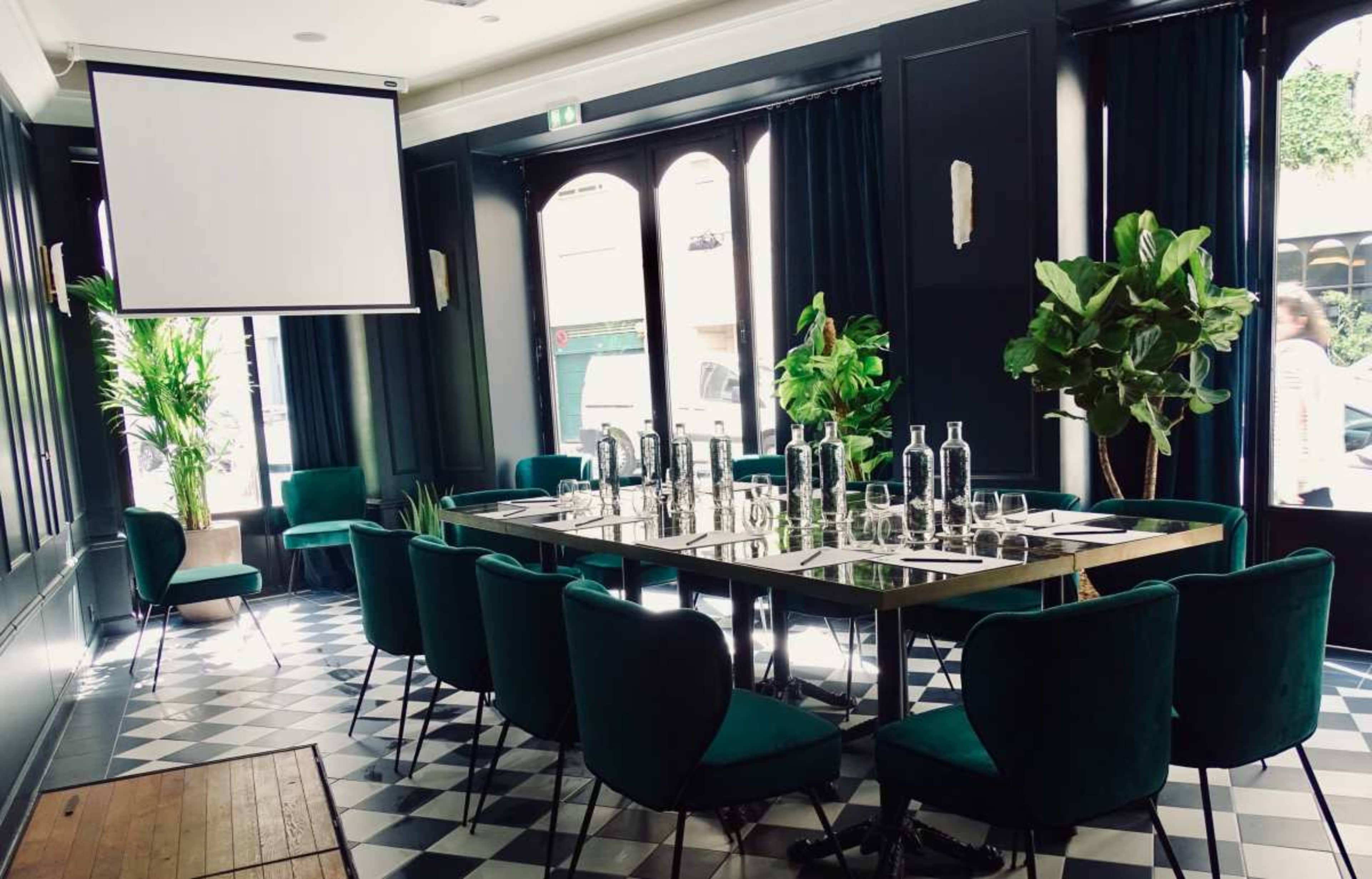 A conference room is set up with a long table surrounded by green chairs, adorned with plants, and featuring a projector screen and a checkered floor.