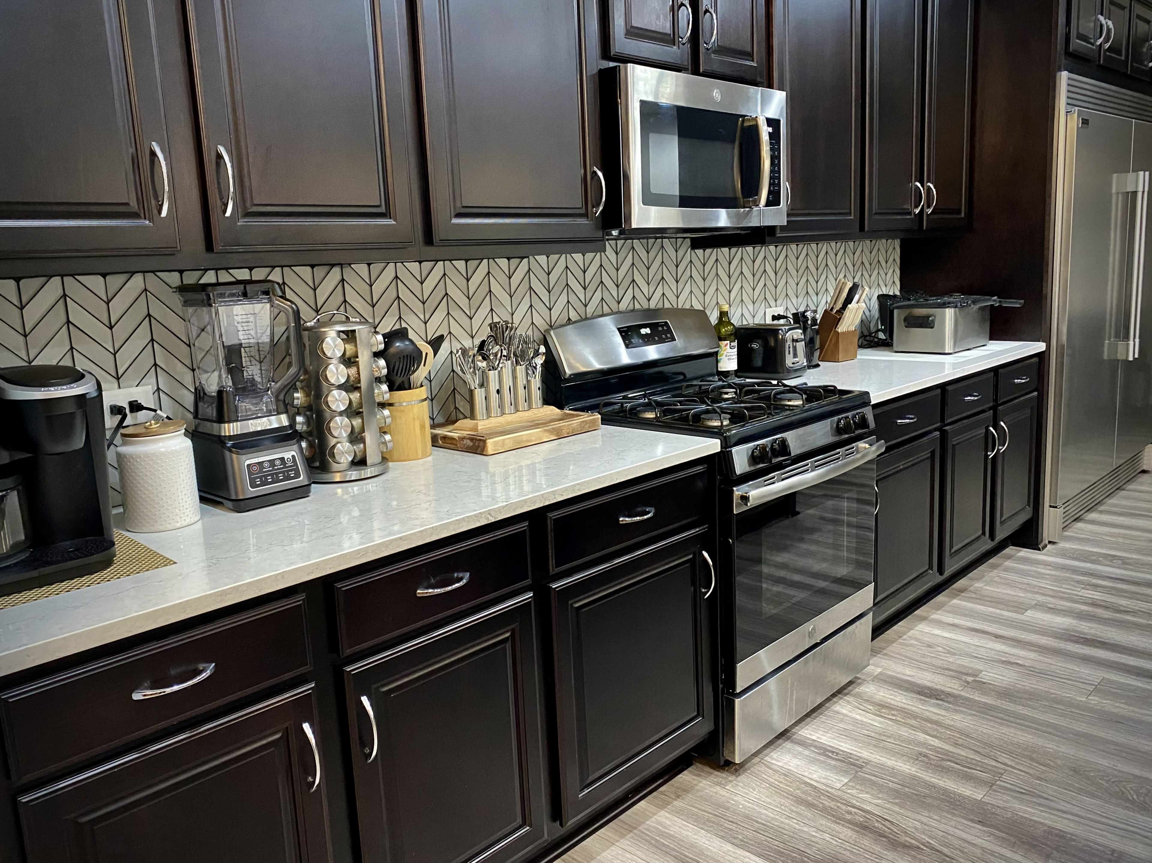 A modern kitchen features dark cabinetry, stainless steel appliances, and a light countertop with various kitchen tools and appliances on display.