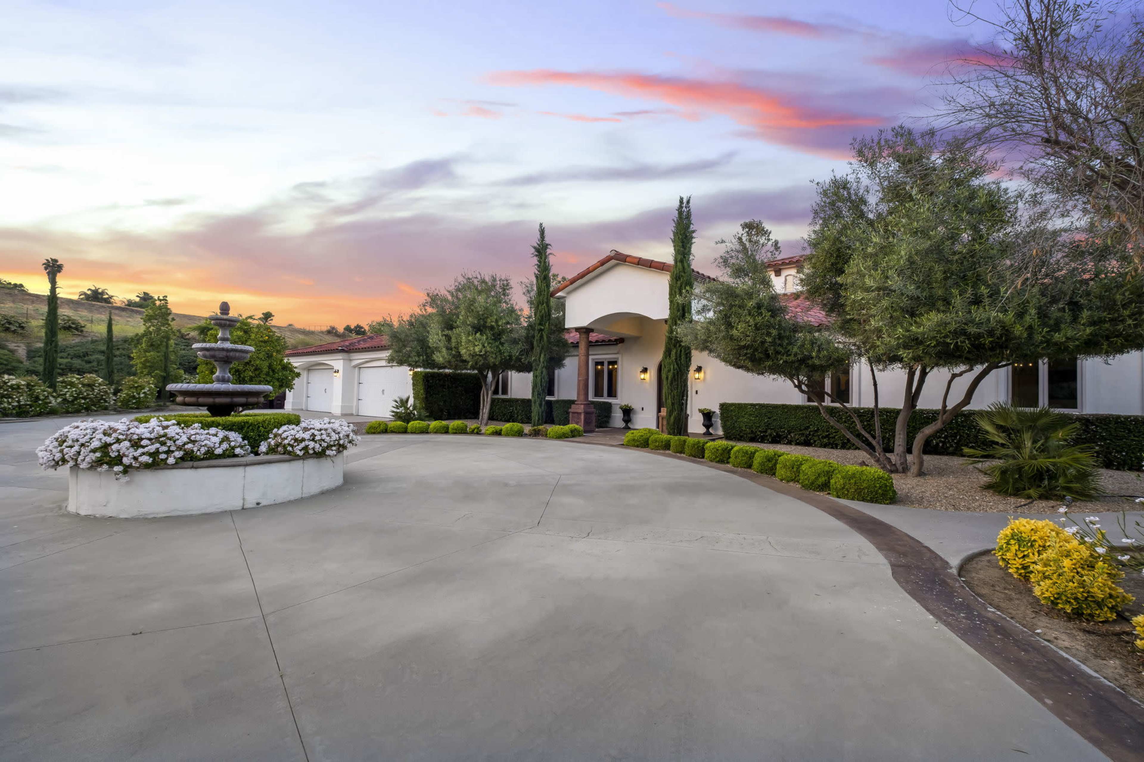 A large, modern house with a fountain and landscaped garden is set against a sunset sky.