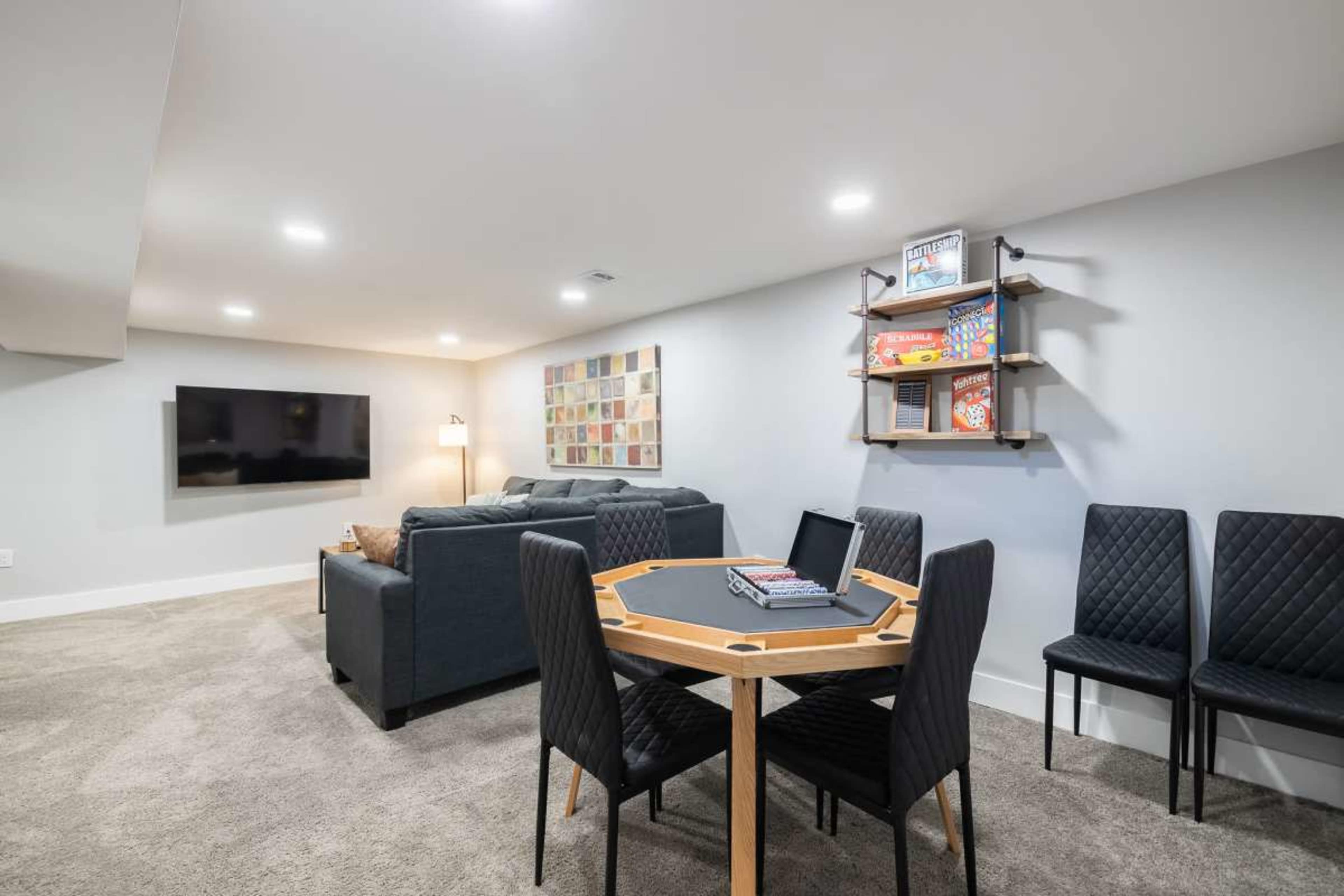 The image shows a modern basement living area featuring a sectional sofa, a wall-mounted TV, a game table with chairs, and a wall shelf displaying books and games.
