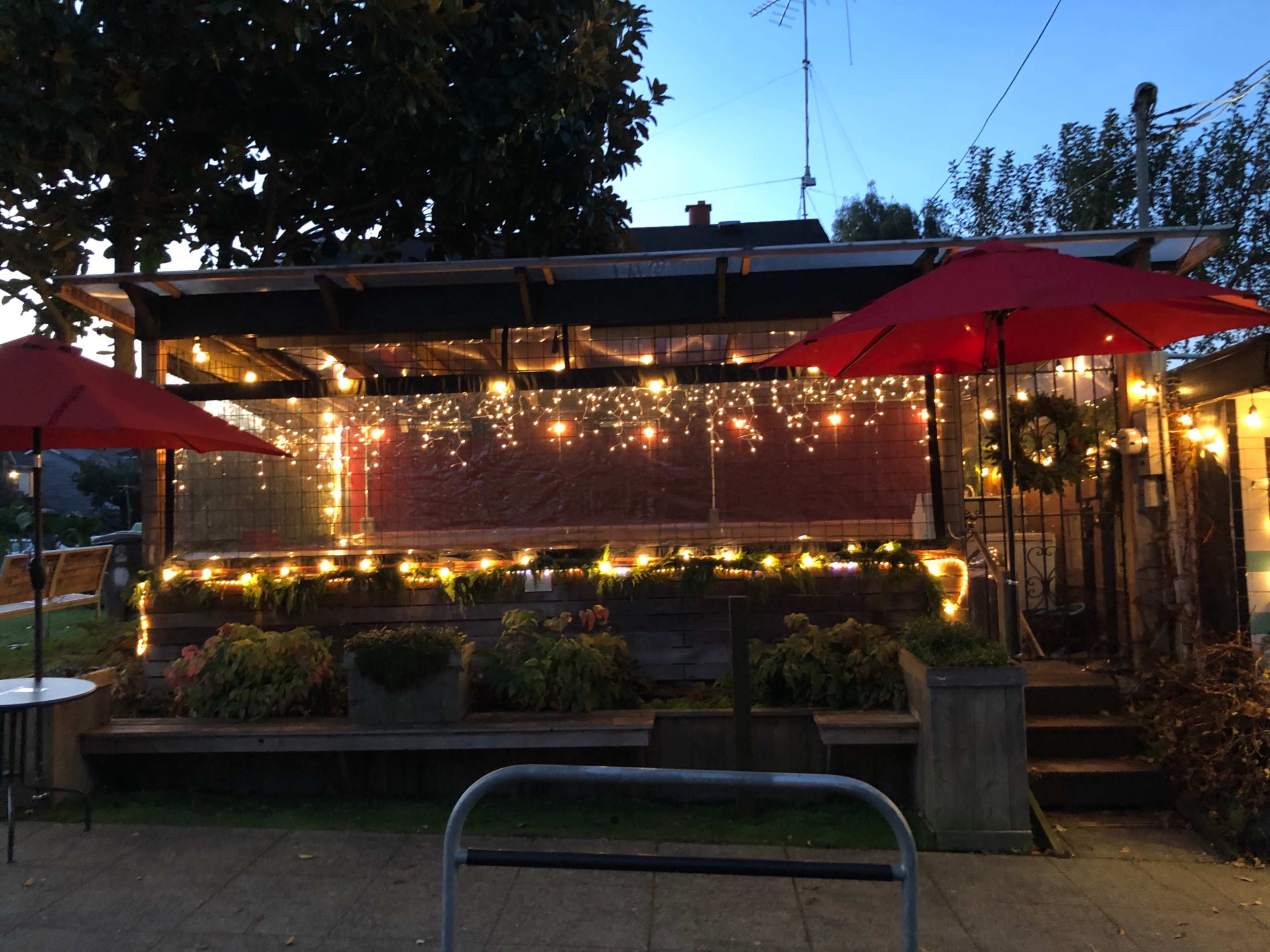 The exterior of a building features outdoor seating under red umbrellas and is adorned with string lights, creating a cozy atmosphere.