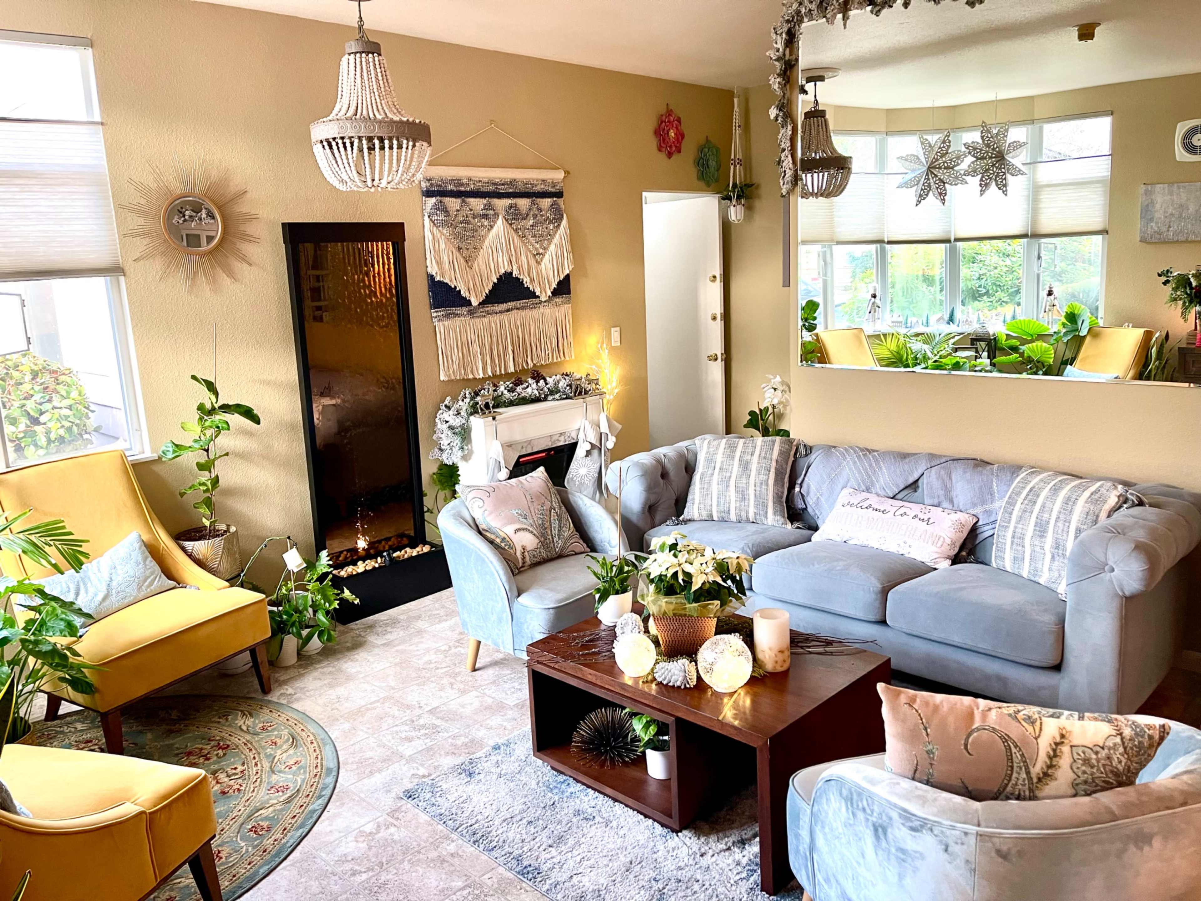 A cozy living room featuring a gray sofa, yellow armchairs, a wooden coffee table, plants, and a decorative fireplace.