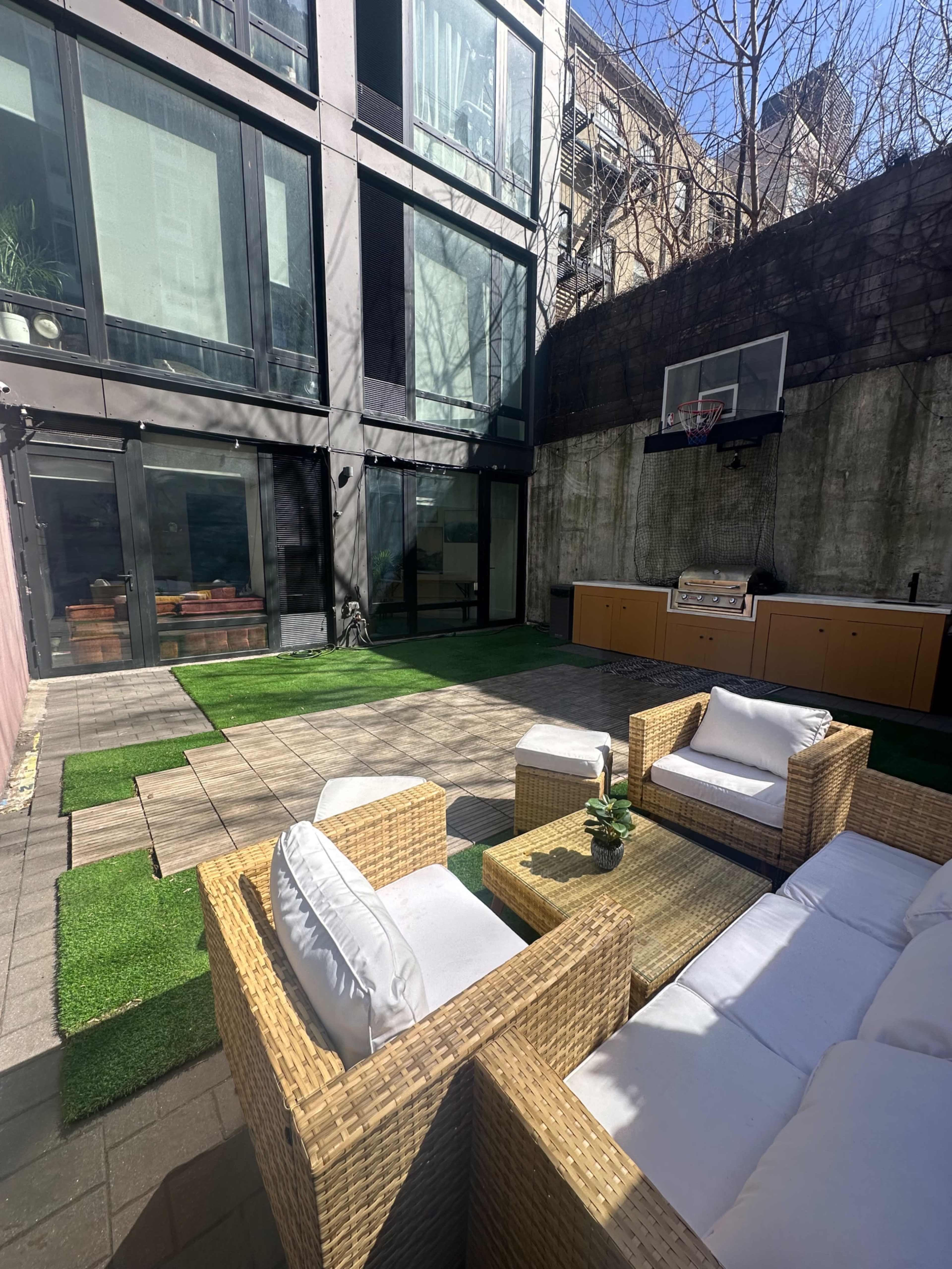 The image shows a modern outdoor patio area featuring wicker furniture, a small table, artificial grass, and a basketball hoop on a wall of a building.