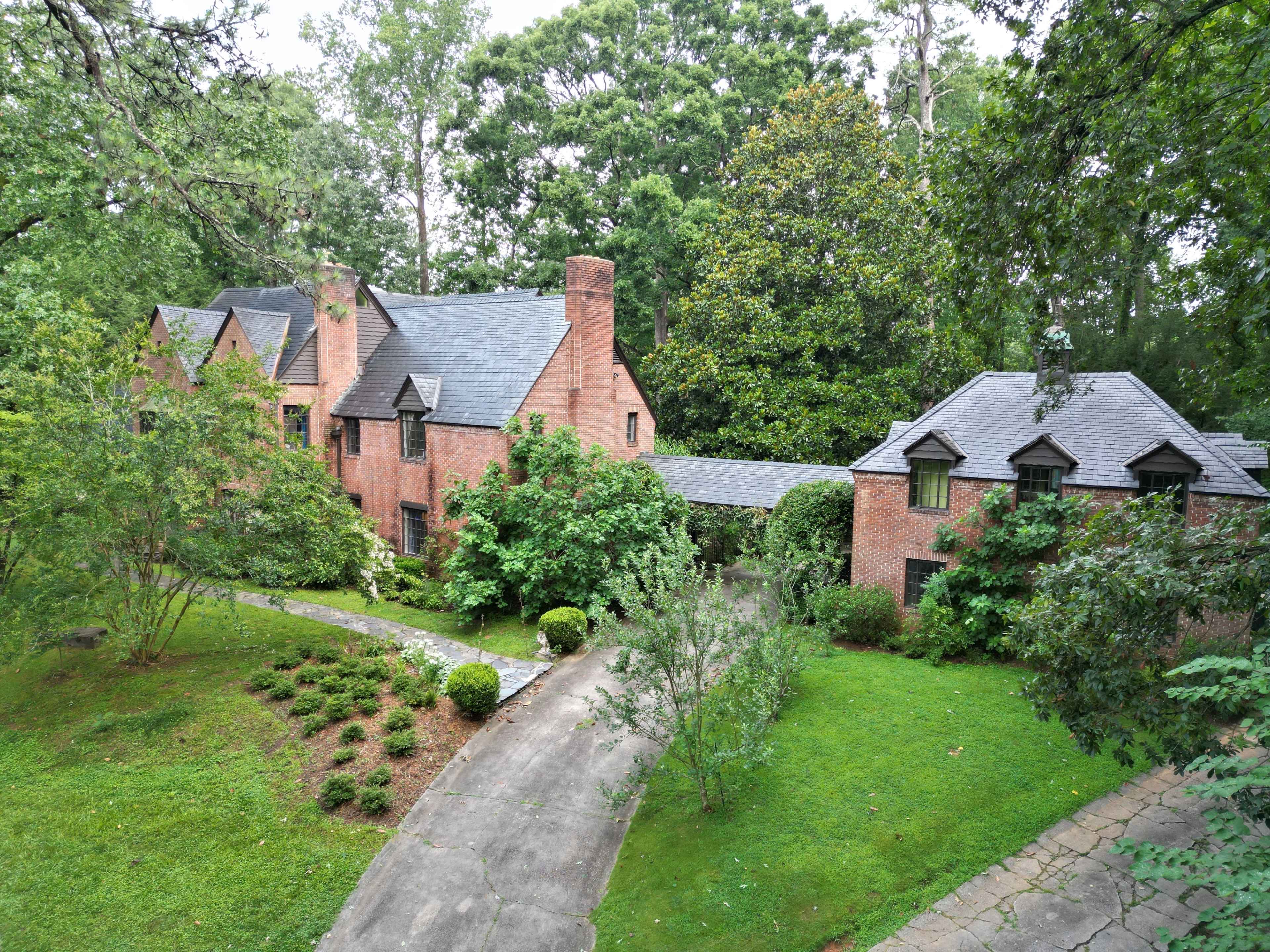 A two-story brick house surrounded by trees and landscaped greenery, with a curved driveway leading up to it.