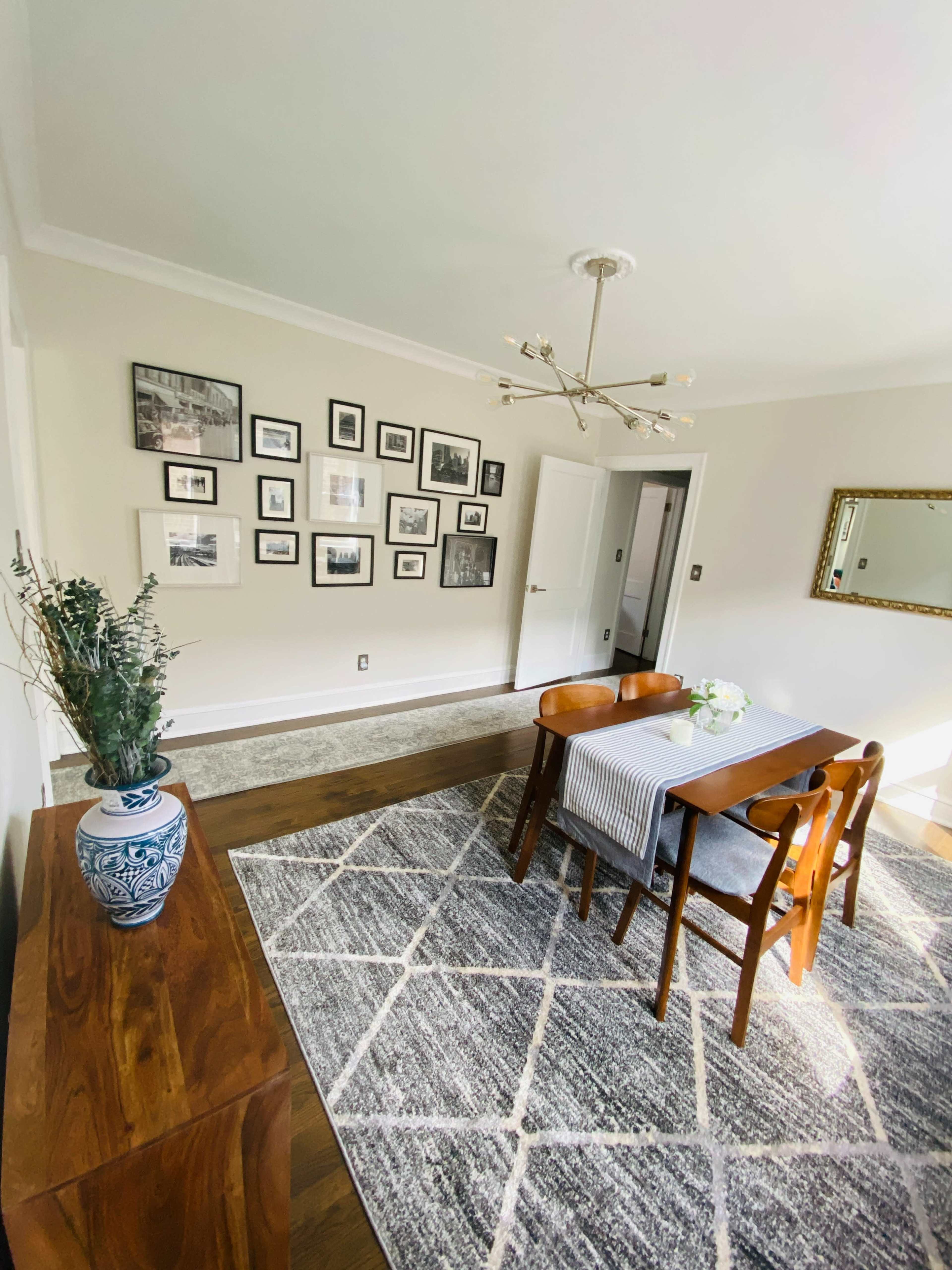 A dining area features a wooden table with chairs, a patterned rug, and a wall adorned with framed pictures.