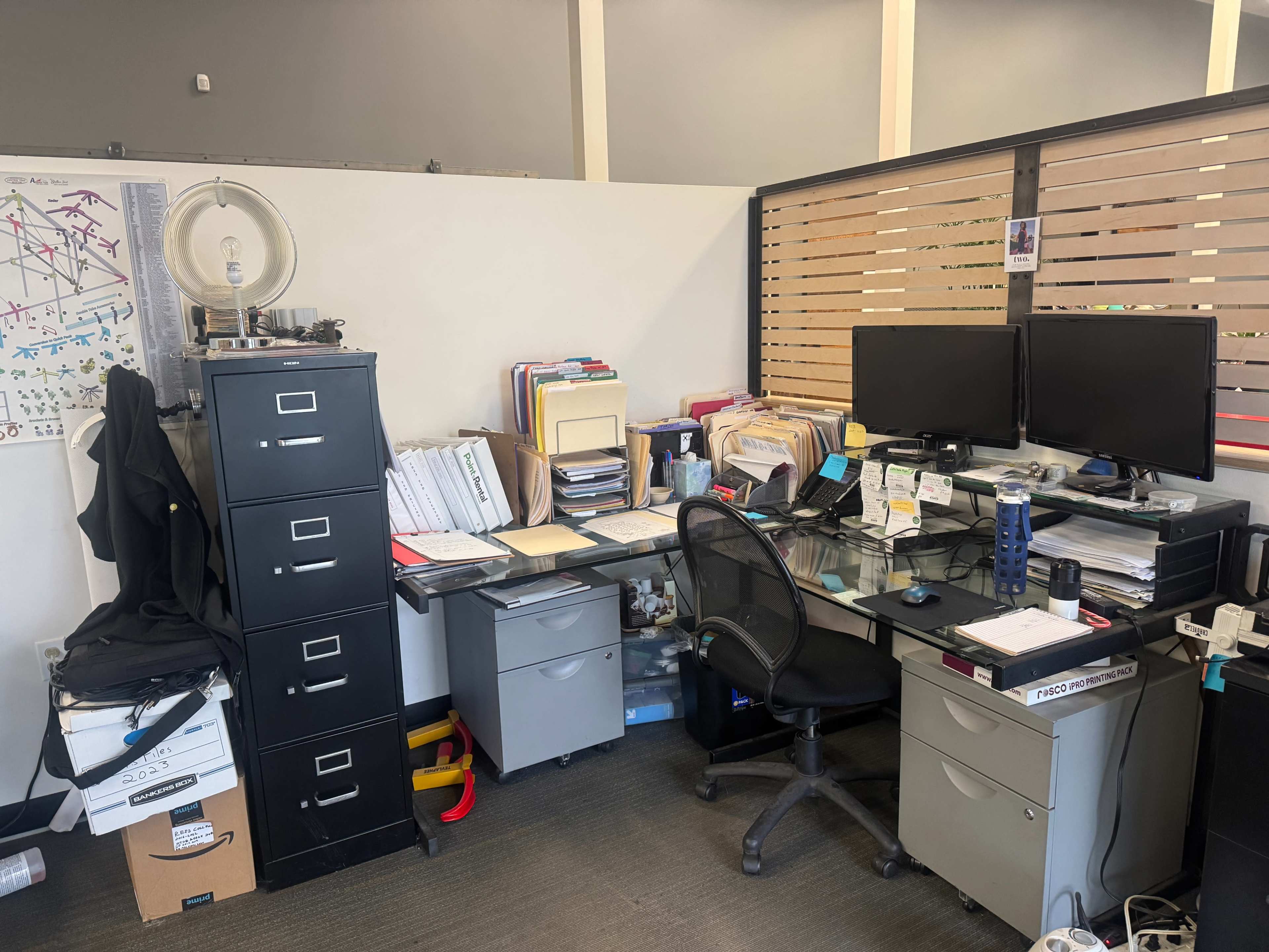 The image shows a cluttered office workspace with two computer monitors, filing cabinets, and stacks of paper on a glass desk.