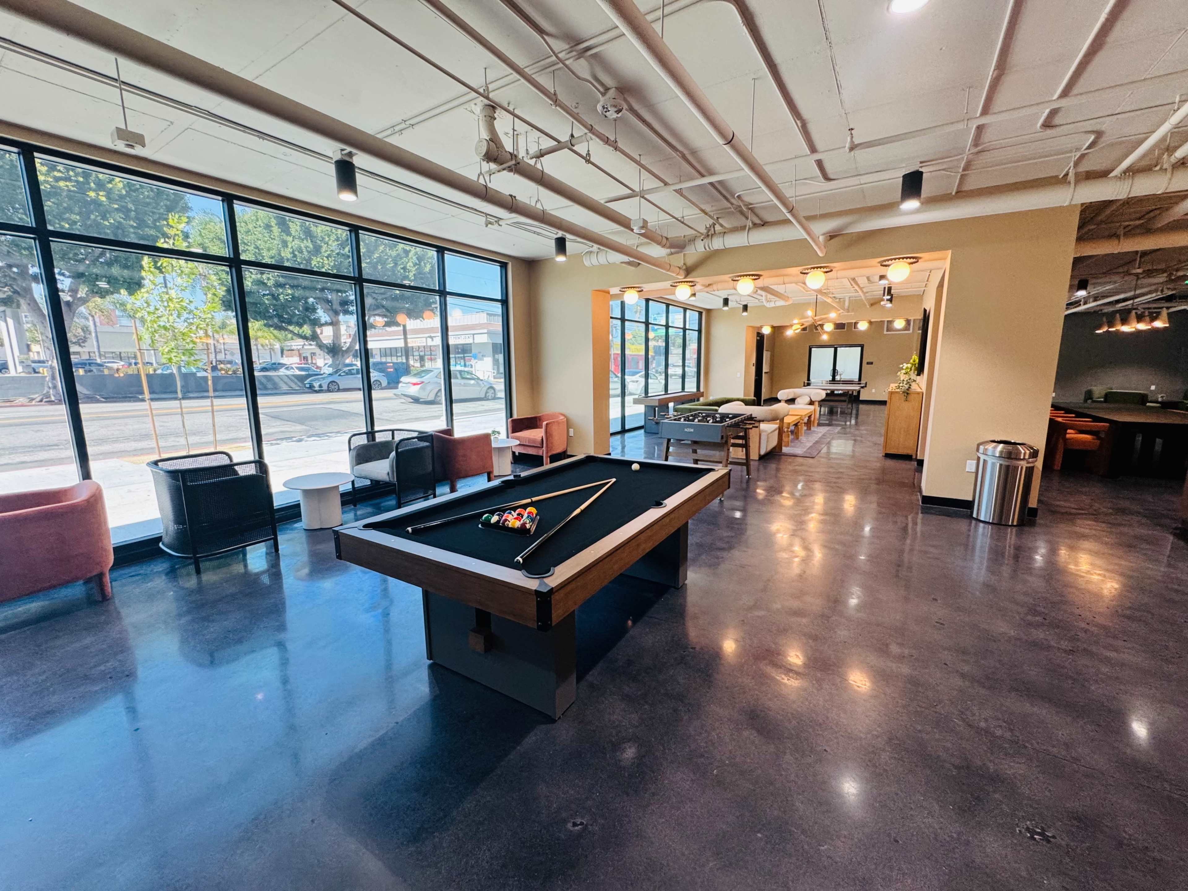 The image shows a spacious and modern interior with a pool table in the center, surrounded by seating areas and large windows letting in natural light.